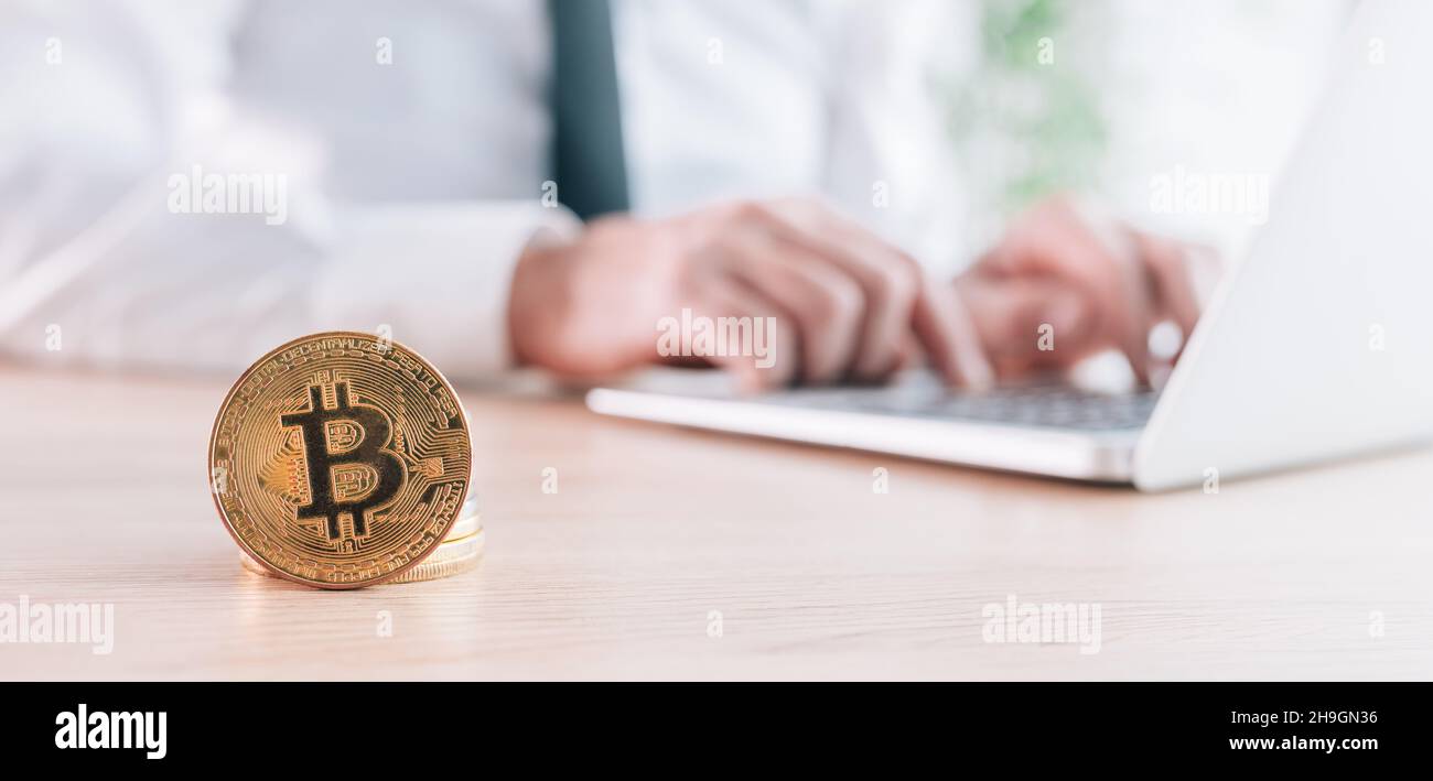 Businessman che commercia con la piattaforma di commercio di criptovaluta del bitcoin sopra il suo calcolatore del laptop in ufficio, monete impilate davanti, immagine panoramica con sel Foto Stock