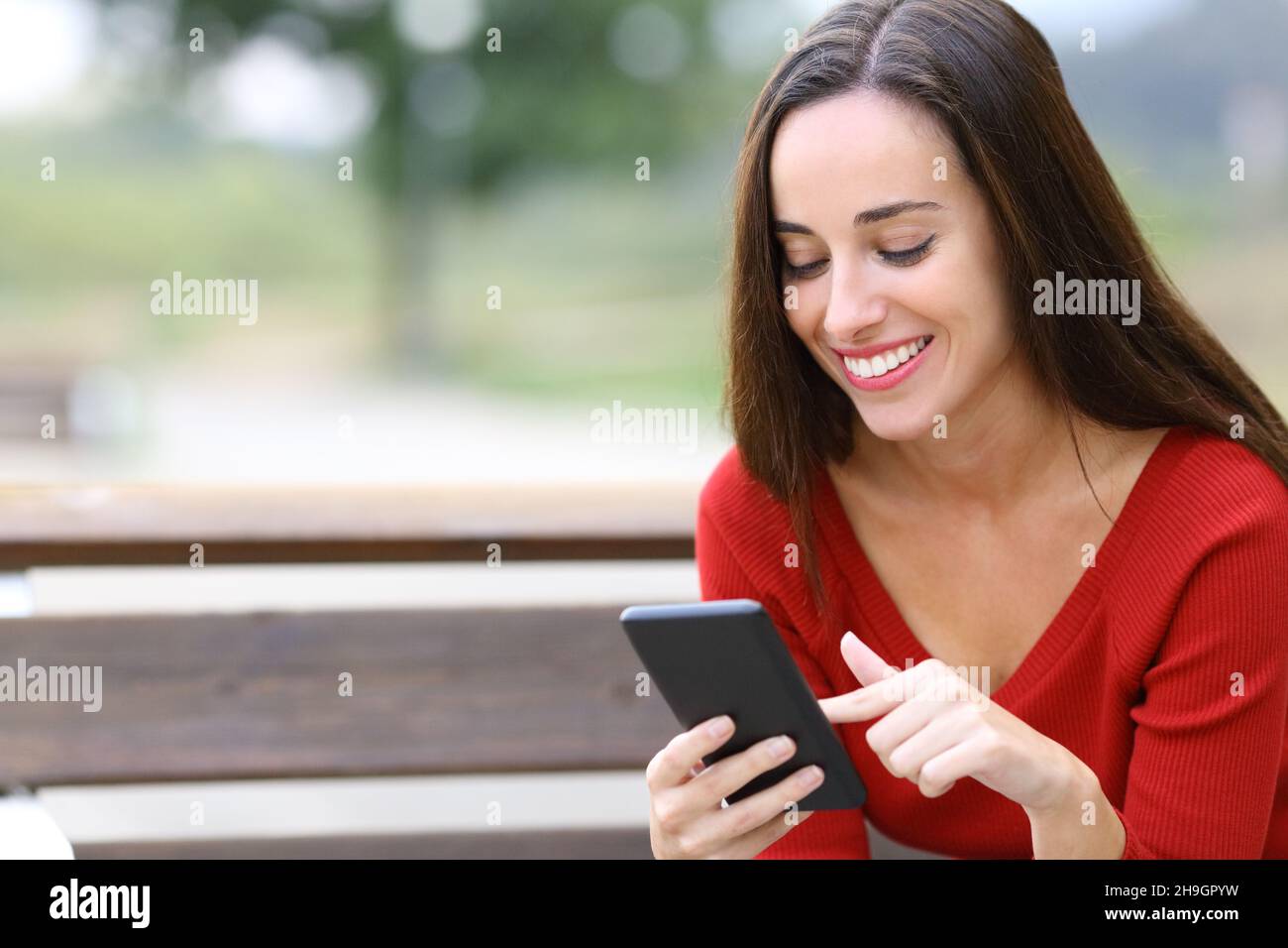Donna felice che naviga contenuti smartphone seduti su una panca in un parco Foto Stock