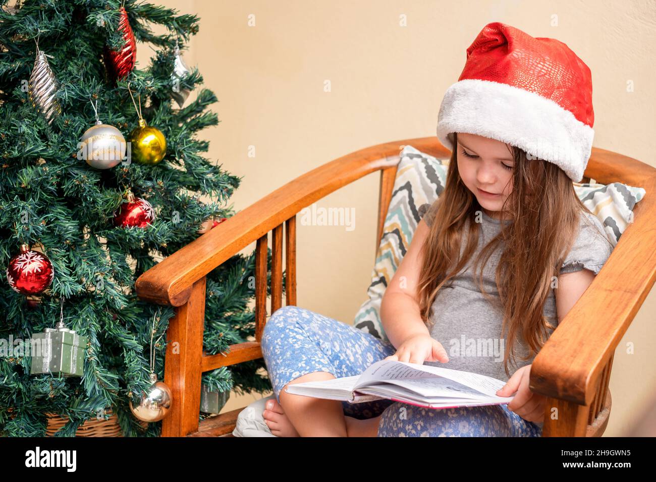 La ragazza siede in un cappello di Santa vicino all'albero di Natale. Un bambino che legge un libro. Natale Foto Stock