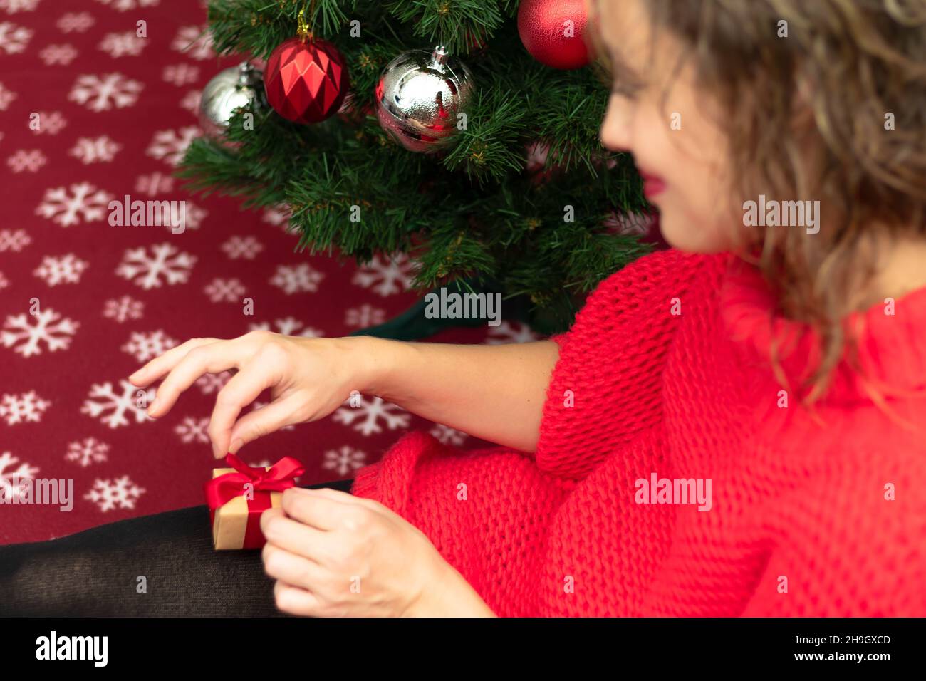 Una bella giovane donna in un maglione rosso tiene una scatola con un regalo legato con un nastro rosso a casa vicino ad un albero di Natale su una coperta calda. Selettivo foc Foto Stock