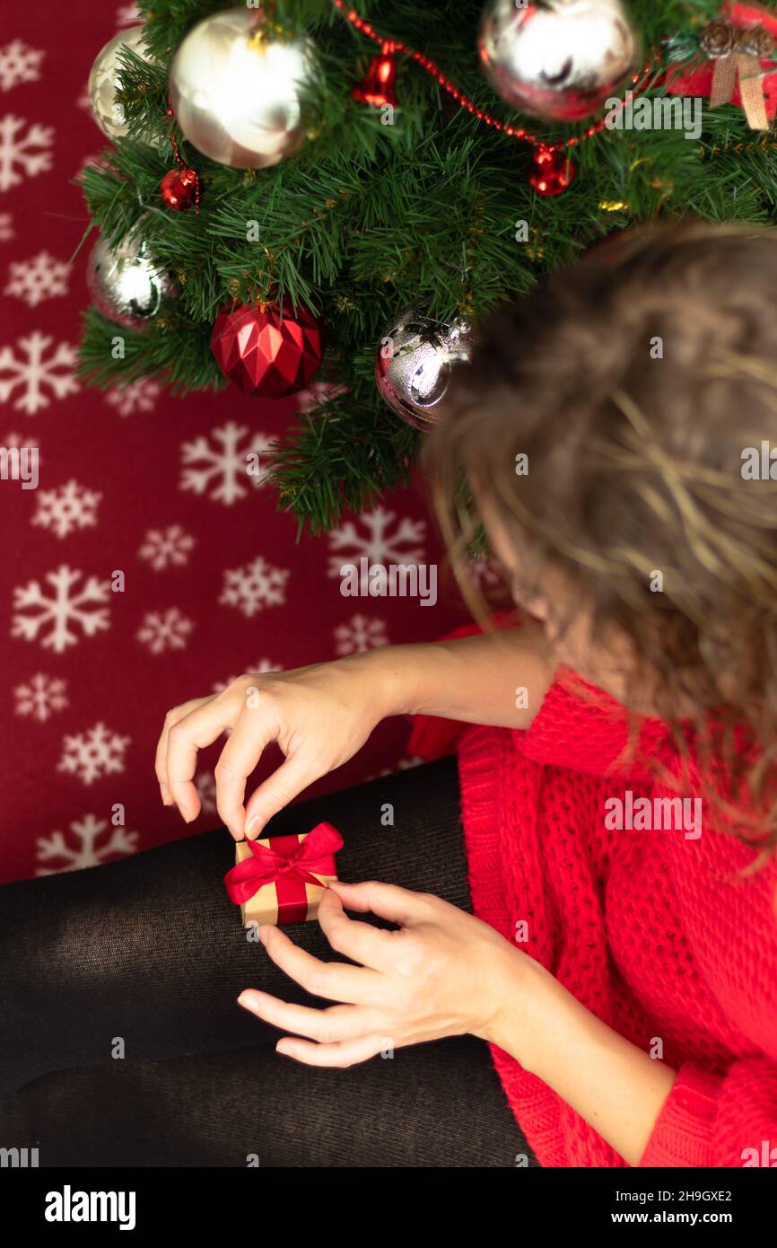 Una bella giovane donna in un maglione rosso tiene una scatola con un regalo legato con un nastro rosso a casa vicino ad un albero di Natale su una coperta calda. Selettivo foc Foto Stock