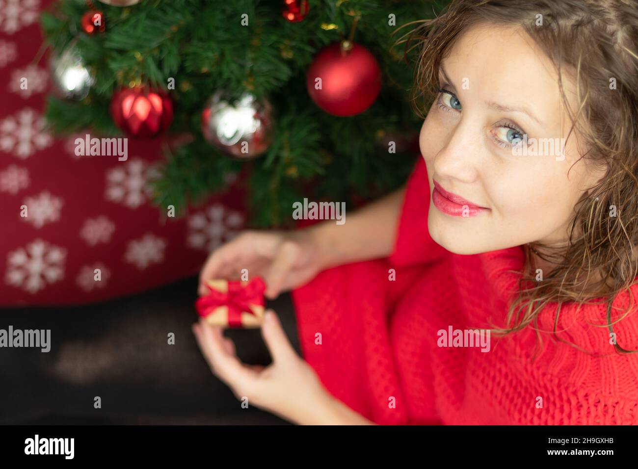 Una bella giovane donna in un maglione rosso tiene una scatola con un regalo legato con un nastro rosso a casa vicino ad un albero di Natale su una coperta calda. Selettivo foc Foto Stock