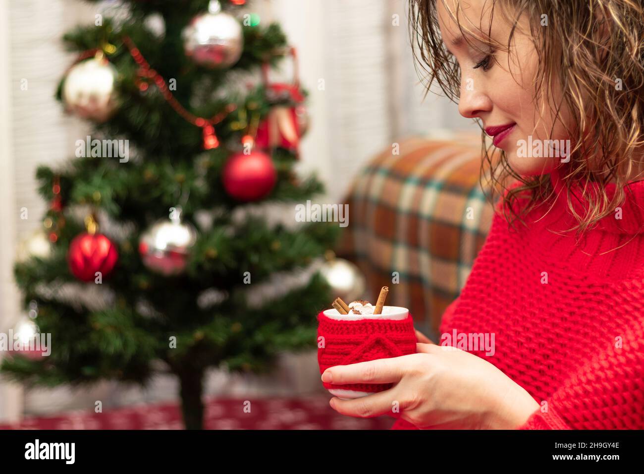 Giovane bella donna in un maglione rosso con una tazza di cacao e marshmallows a casa dall'albero del nuovo anno alla vigilia di Capodanno. Messa a fuoco selettiva. Verticale Foto Stock