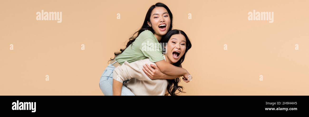 stupito madre asiatica piggybacking figlia adulta con bocca aperta isolato su beige, banner Foto Stock