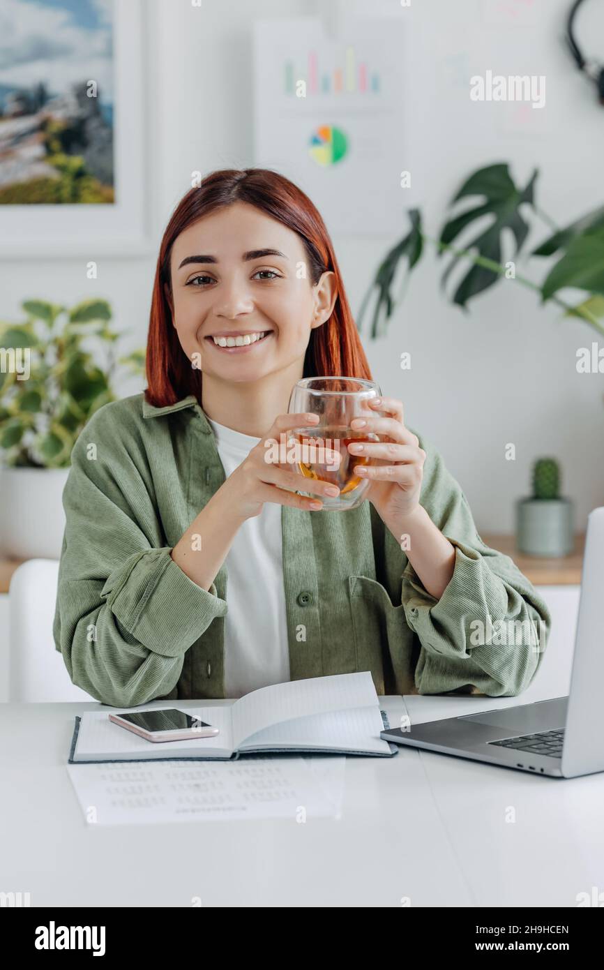 Ritratto di una giovane donna che lavora a casa con un computer portatile. Donna d'affari rilassata che tiene una tazza di tè. Concetto di business o comunicazione online, freelance, Foto Stock