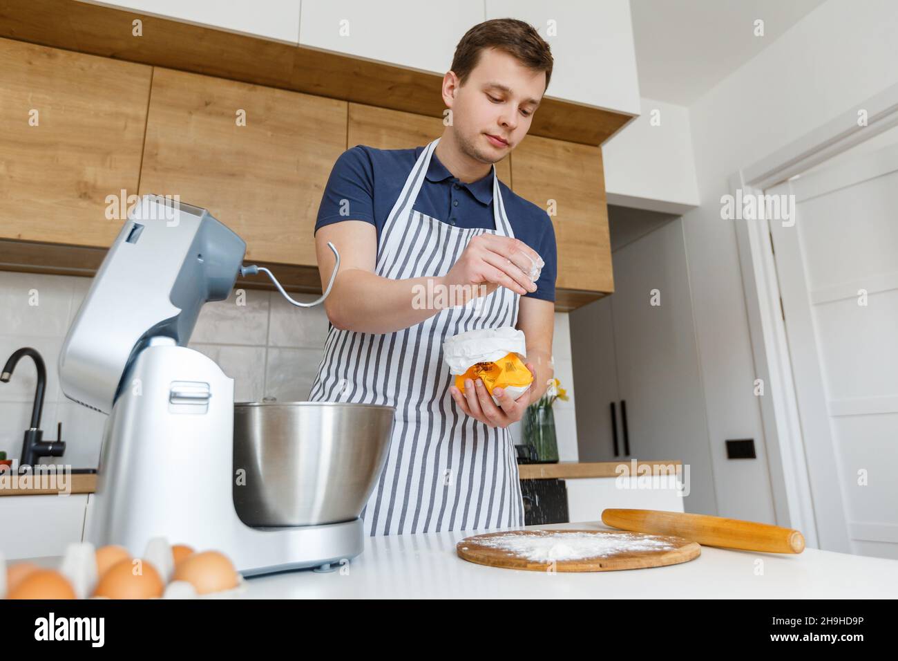 Giovane bell'uomo in grembiule setacciando la farina in cucina moderna. Concetto di prodotti da forno fatti in casa, cucina maschile e stile di vita domestico. Chef pasticceria Foto Stock