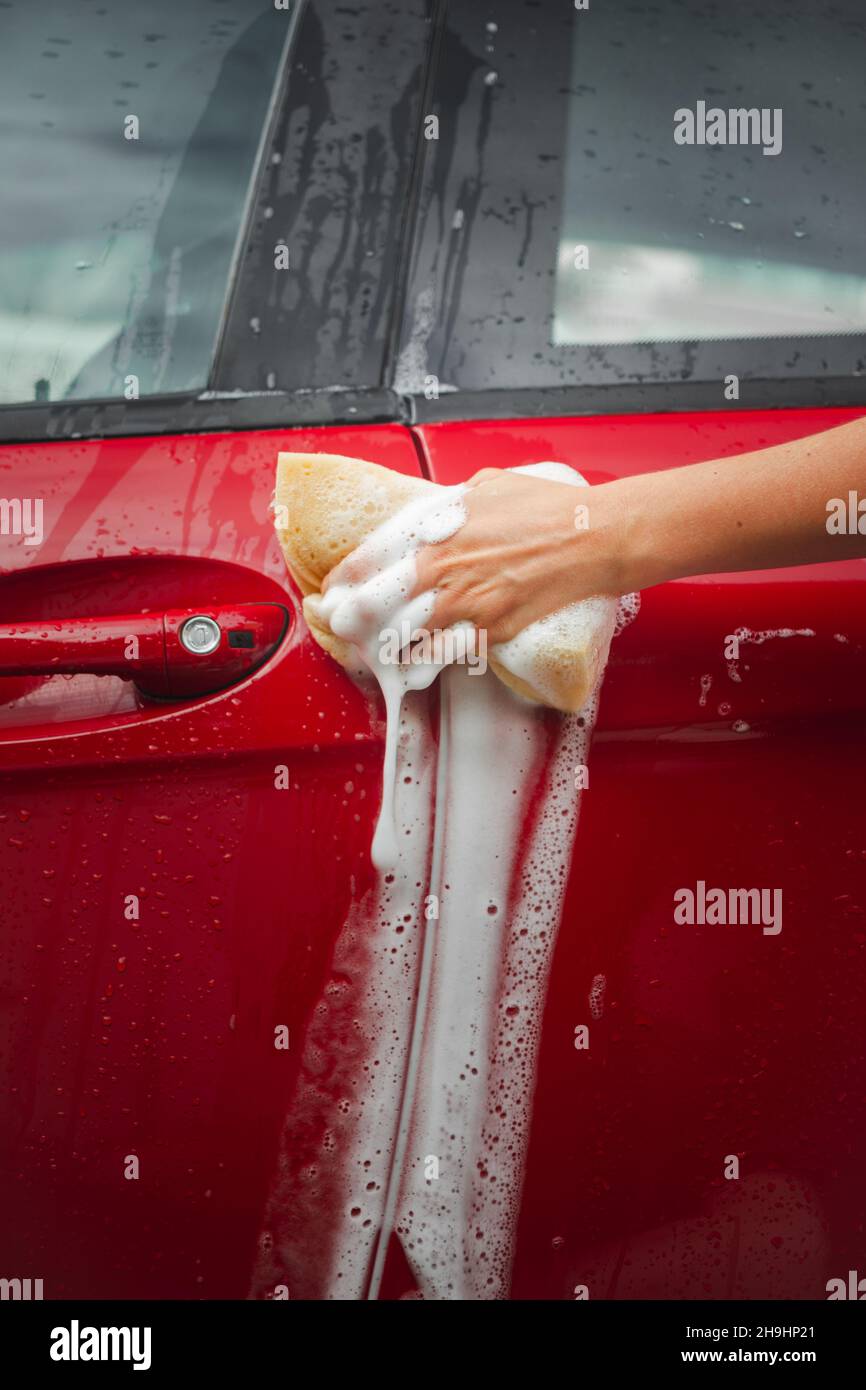 Mano che stringe una spugna con sapone su una vettura della porta, piena di gocce d'acqua. Foto Stock