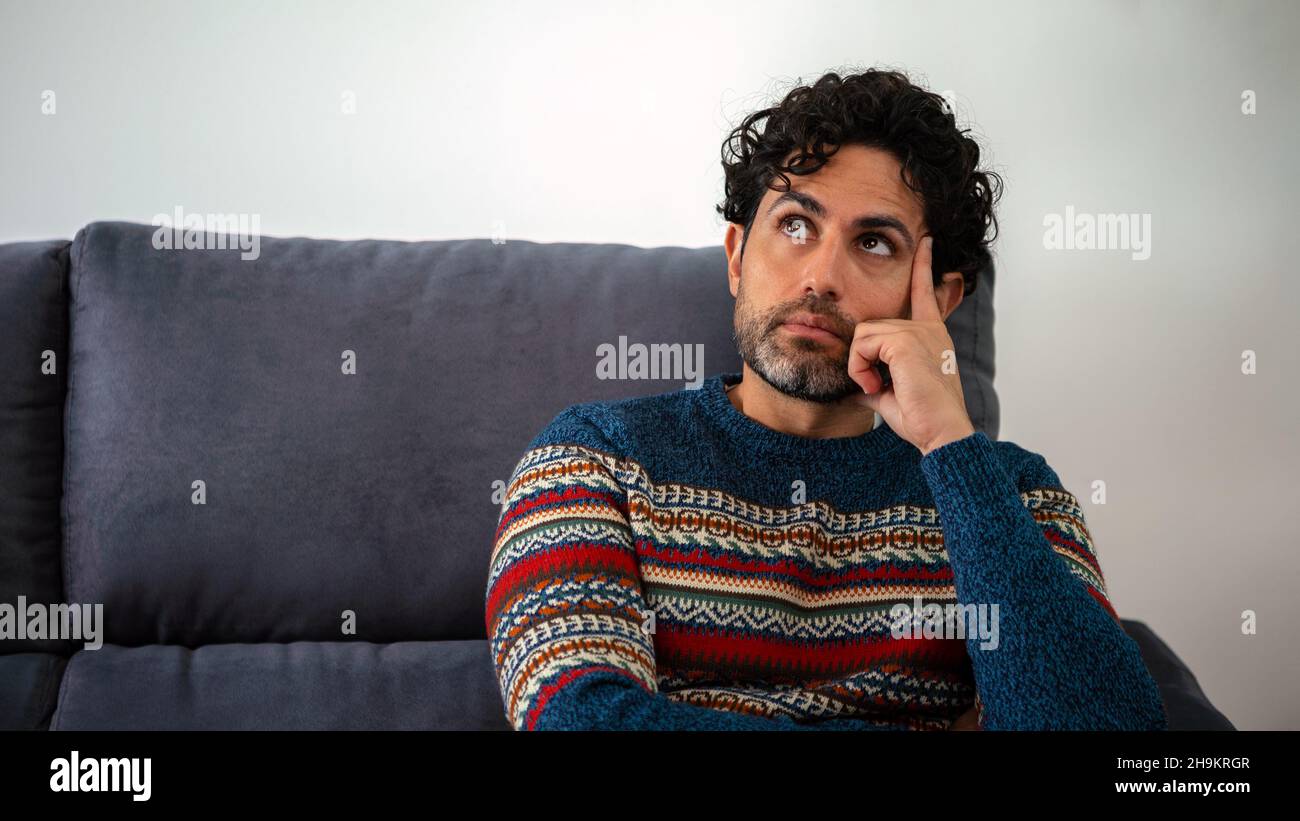 Ritratto di bell'uomo che indossa un maglione invernale e pensare provare a risolvere il problema posa su un divano a casa interno. L'uomo caucasico preoccupato seduto così Foto Stock