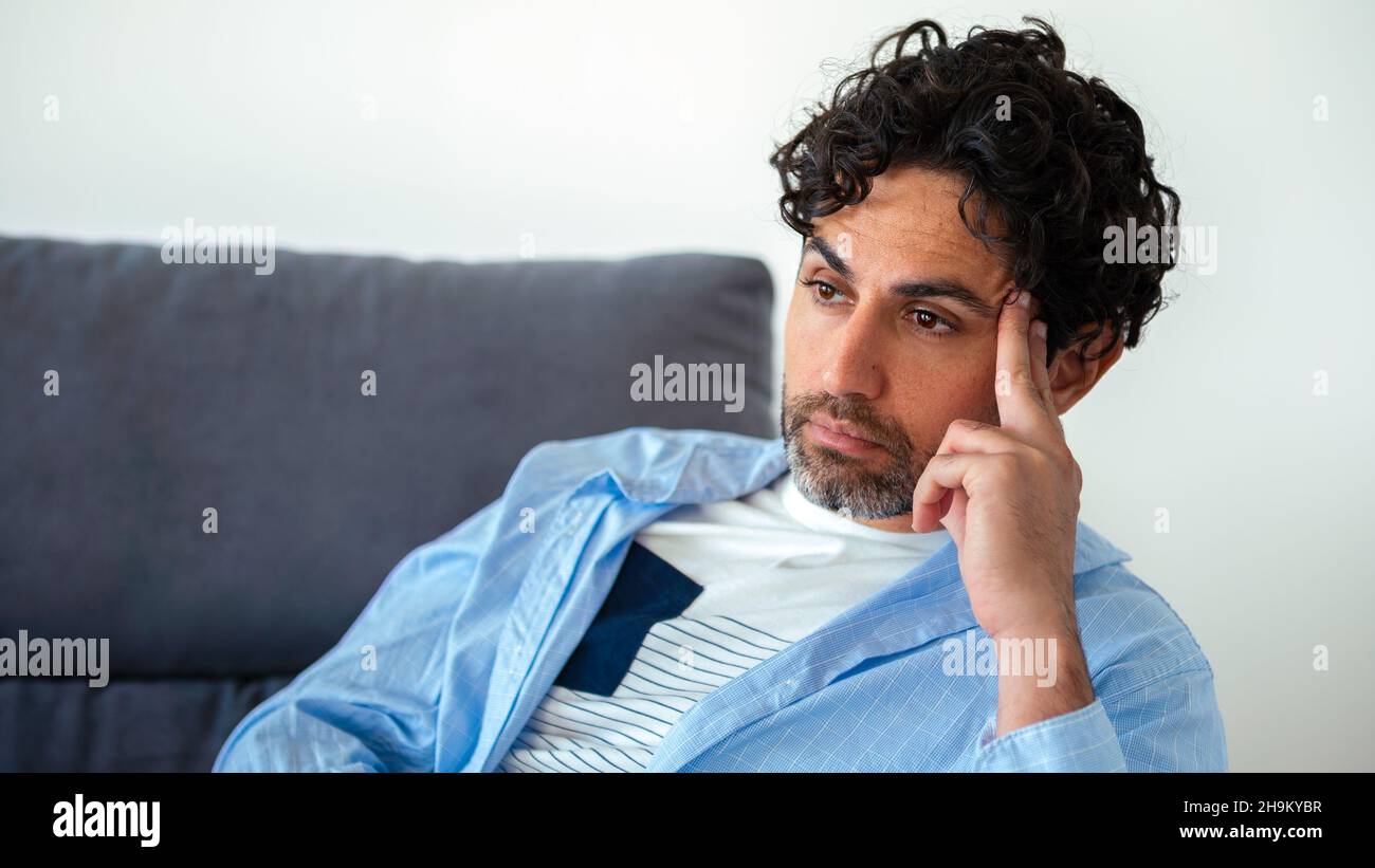 Ritratto di bell'uomo che indossa una camicia e pensare provare a risolvere il problema posa su un divano a casa interno. Uomo caucasico preoccupato seduto sul divano di hou Foto Stock