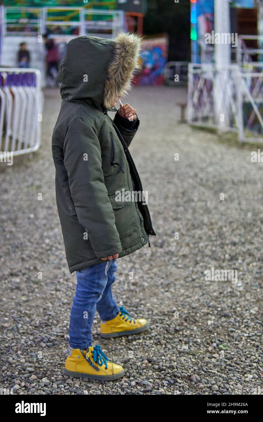 bambino latino vestito di jeans e parka verde in pelliccia cappuccio perso in un parco di divertimento. inverno, verticale Foto Stock