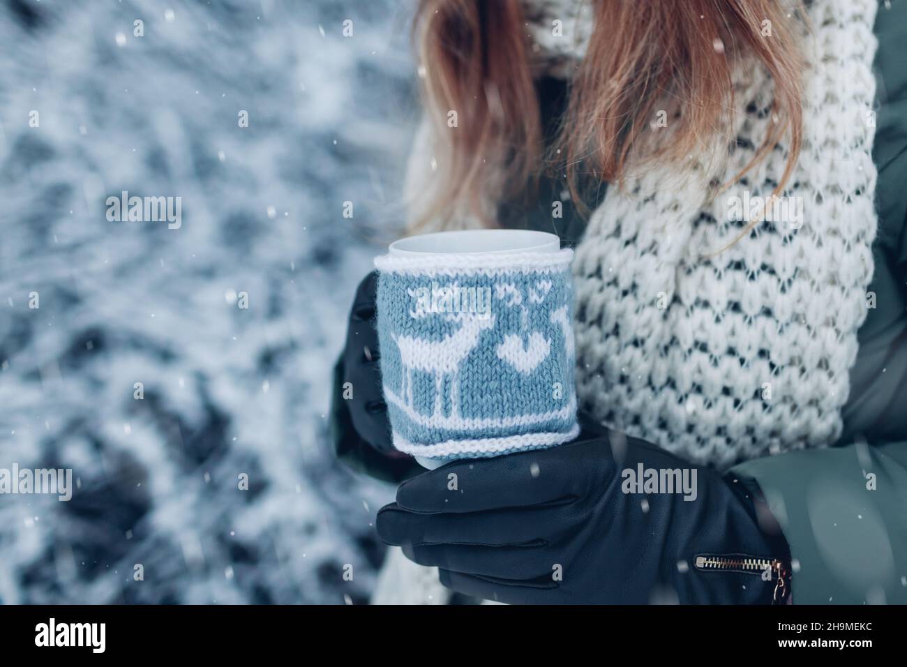 Primo piano di una tazza di tè o caffè vestito con cappotto di Natale a maglia in mano femminile nel parco invernale. Bevande per riscaldarsi in condizioni di clima ghiacciato all'aperto Foto Stock