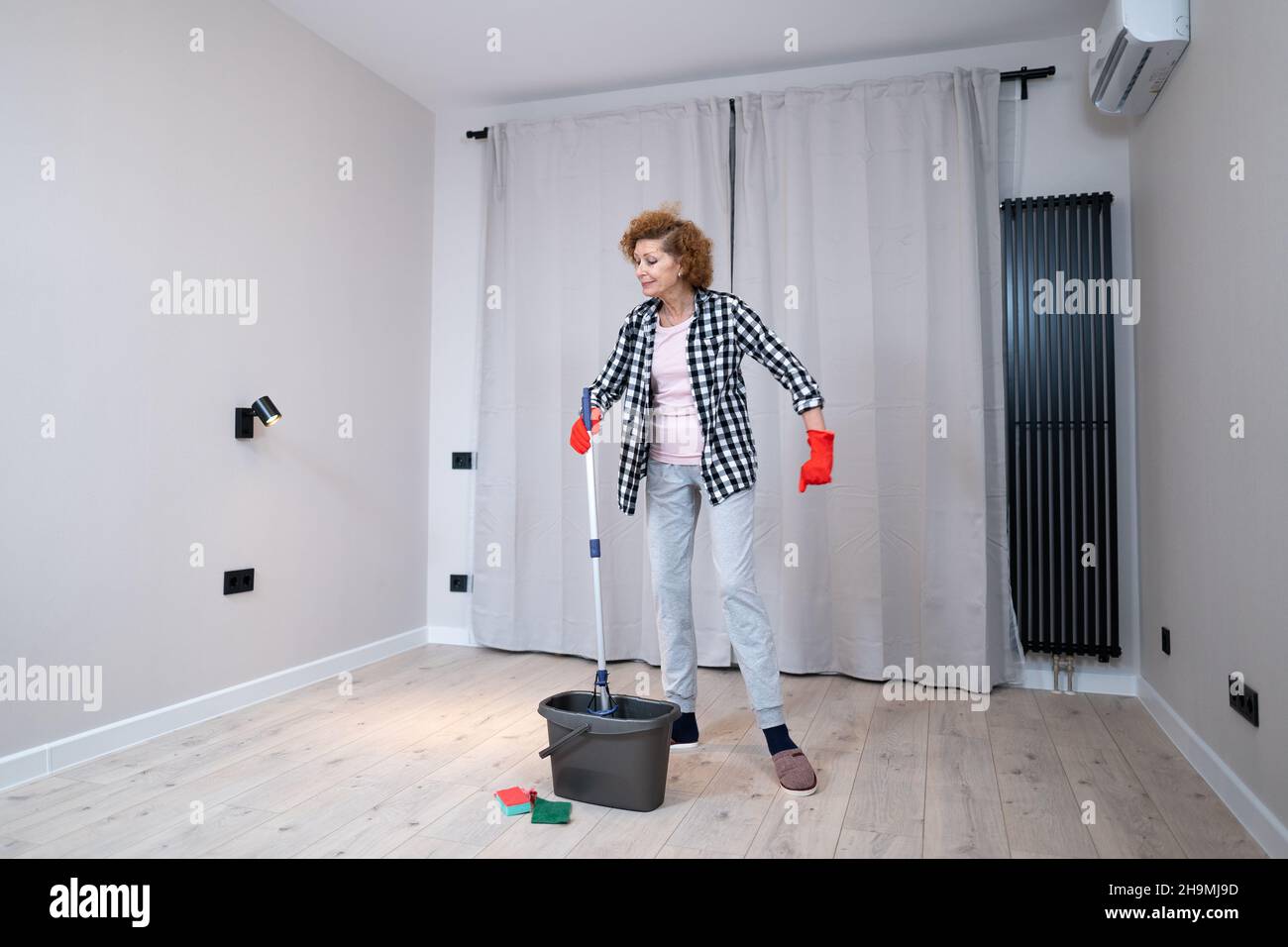 Persone, lavori domestici e concetto di pulizia. Donna anziana felice in guanti protettivi pulisce il pavimento e danze a casa in appartamento vuoto prima di muoversi Foto Stock