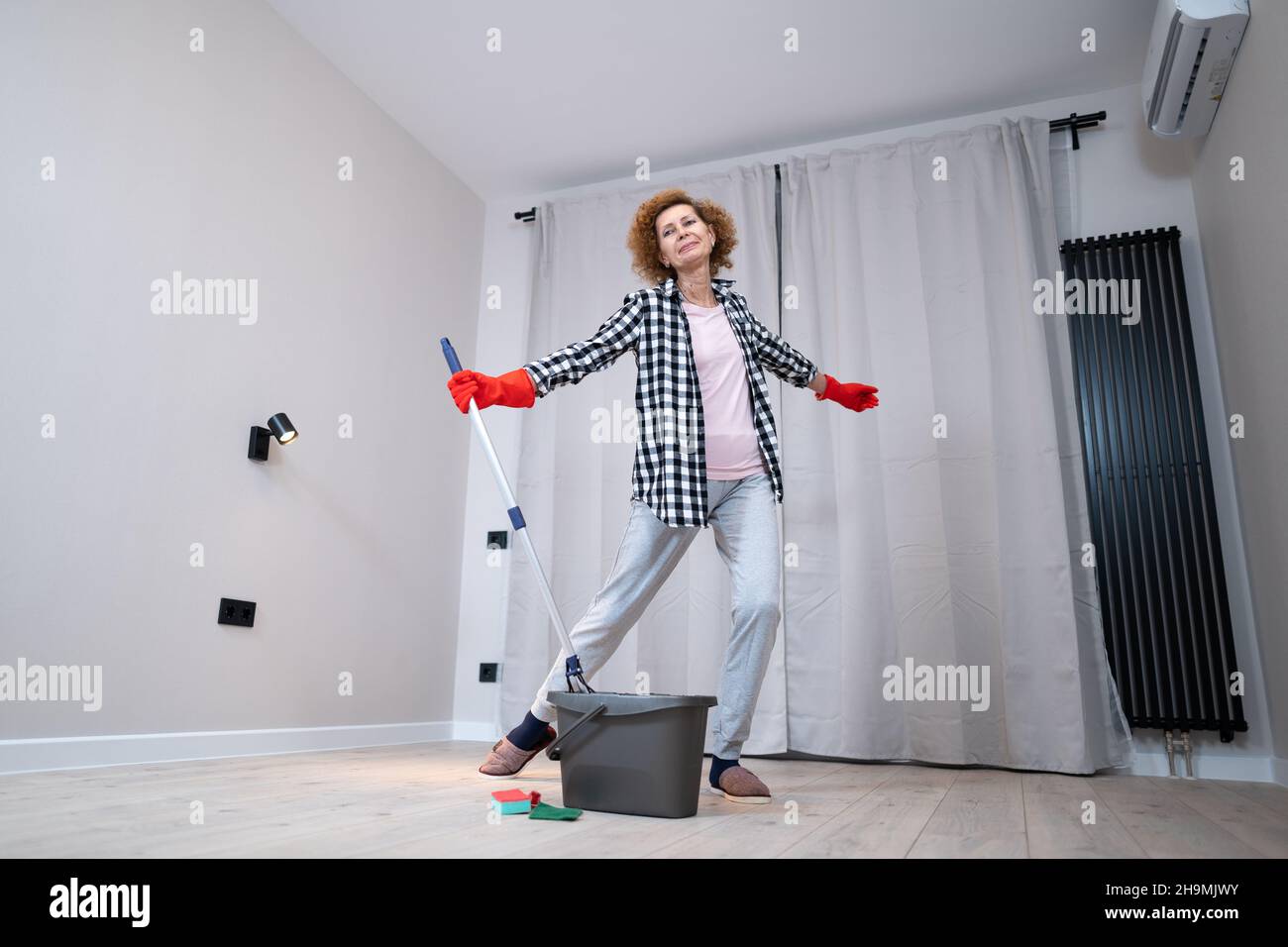 Persone, lavori domestici e concetto di pulizia. Donna anziana felice in guanti protettivi pulisce il pavimento e danze a casa in appartamento vuoto prima di muoversi Foto Stock