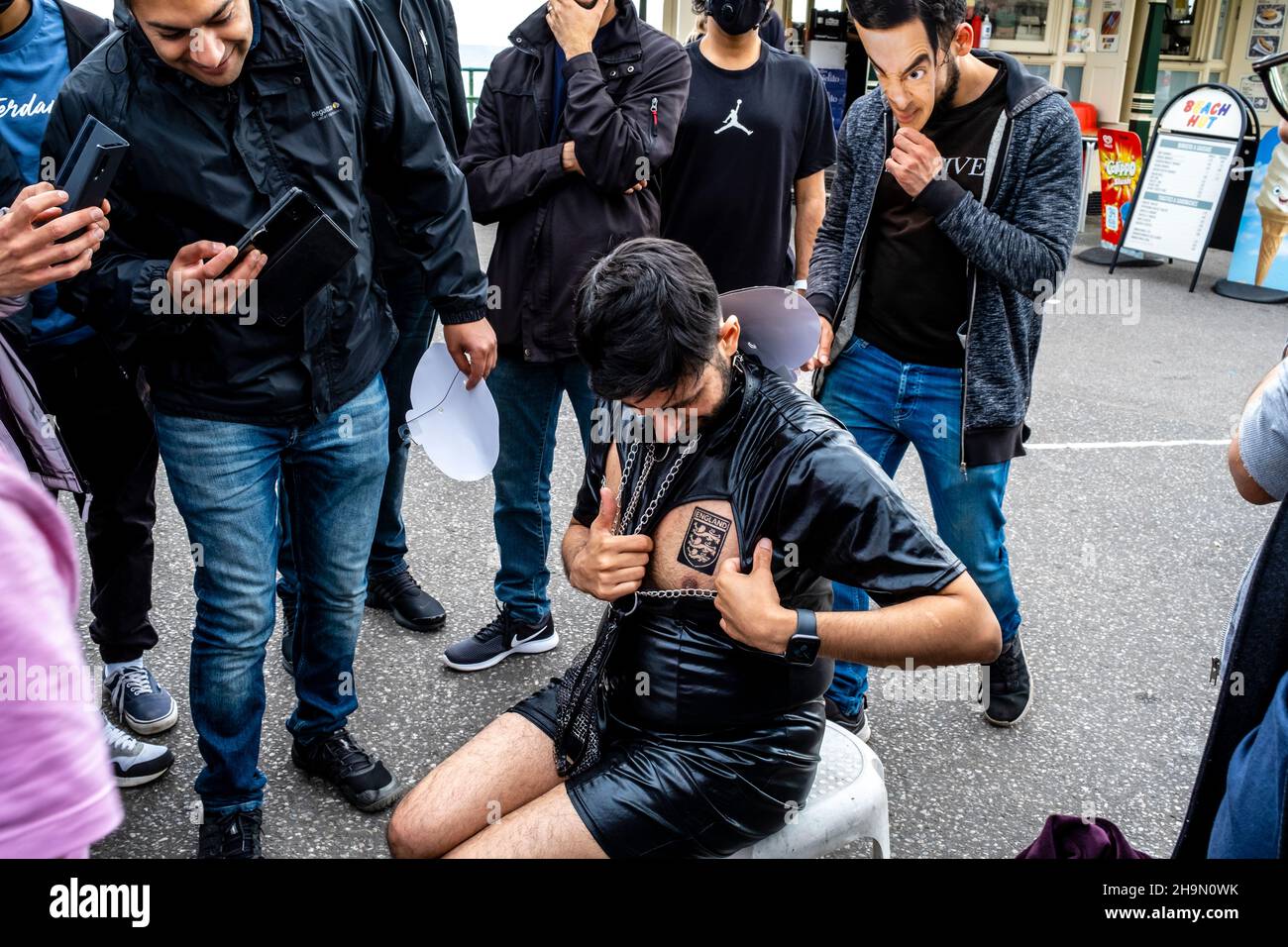 Un gruppo di visitatori britannici/asiatici guarda come uno dei loro amici ha Un tatuaggio temporaneo messo sul suo Chest, Brighton, Sussex, Regno Unito. Foto Stock