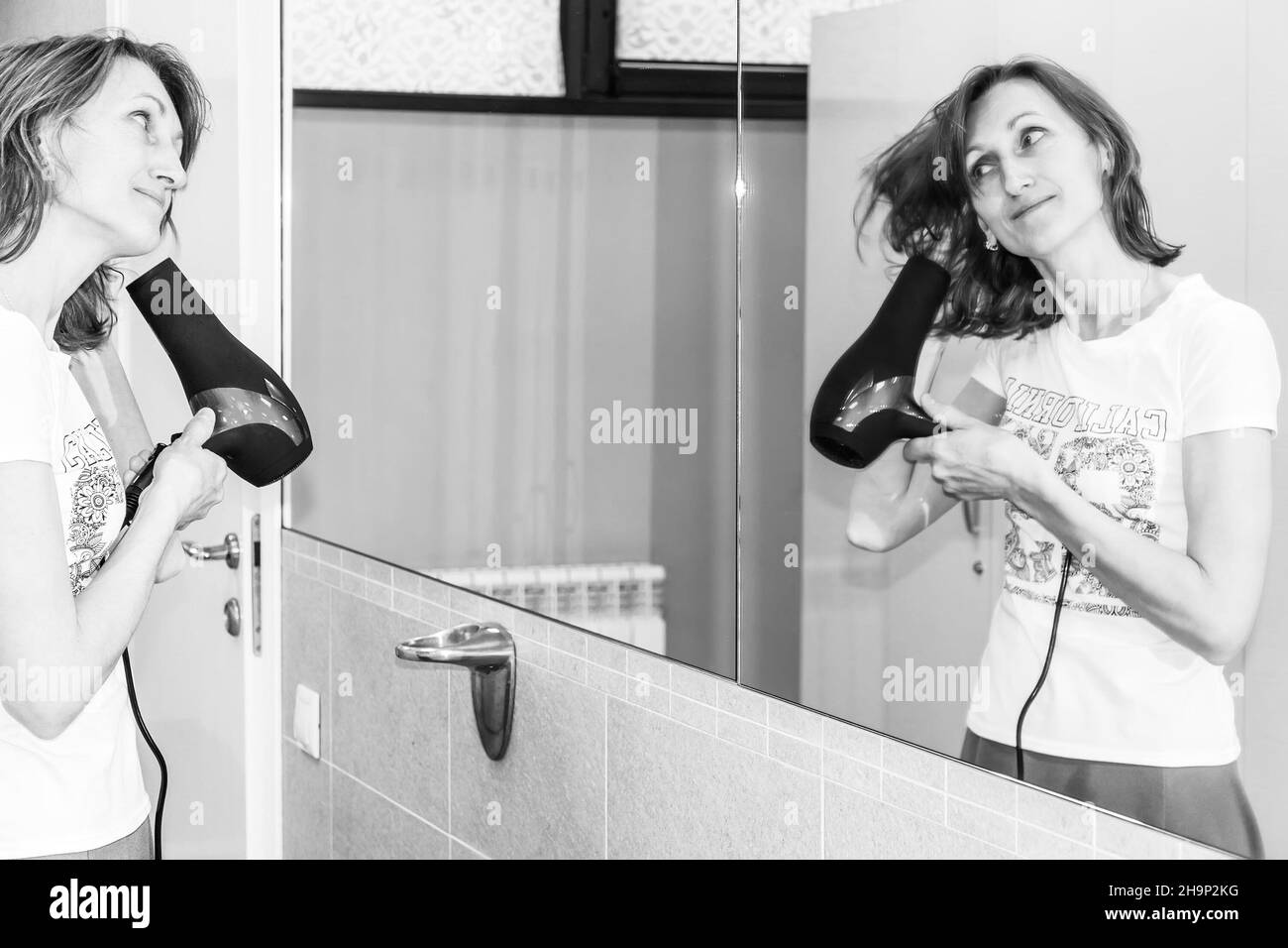 la giovane donna asciuga i capelli con un asciugacapelli davanti a una foto in bianco e nero a specchio Foto Stock