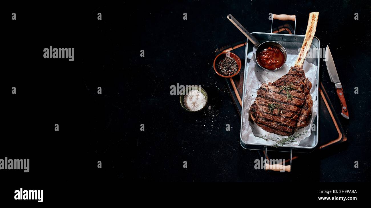Da sopra di deliziosa bistecca di tomahawk alla griglia servita con gustose salse su vassoio di metallo posto vicino al coltello sul tavolo nero Foto Stock