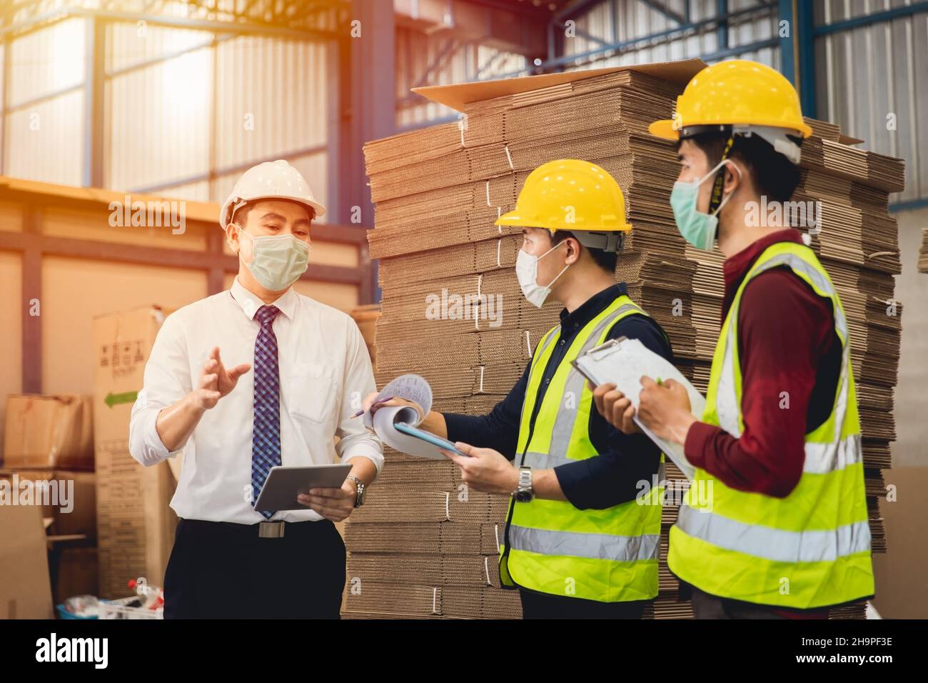 Il responsabile del magazzino dello stabilimento parla con un operatore asiatico che lavora insieme con la maschera di sicurezza e facciale Foto Stock