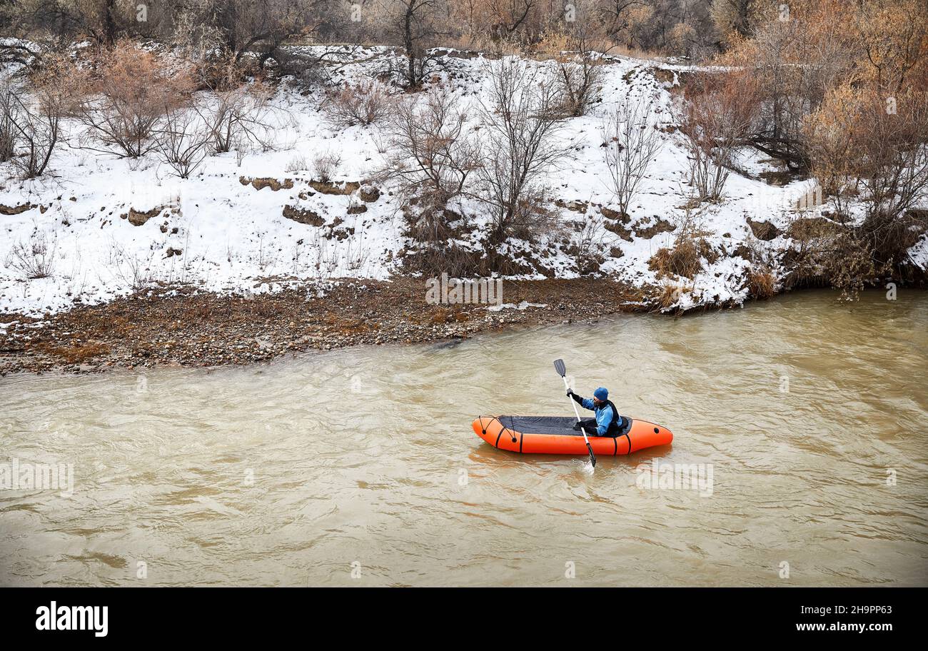 L'uomo dilettante con la barba è pagaiare su barca di zattera arancione nel fiume di acqua sporca in inverno Foto Stock