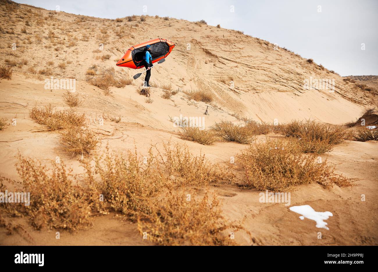 Vecchio uomo con barba nera in giacca blu che tiene barca sportiva arancione zattera alla duna di sabbia sullo sfondo del deserto. Avventura e concetto di viaggio. Foto Stock