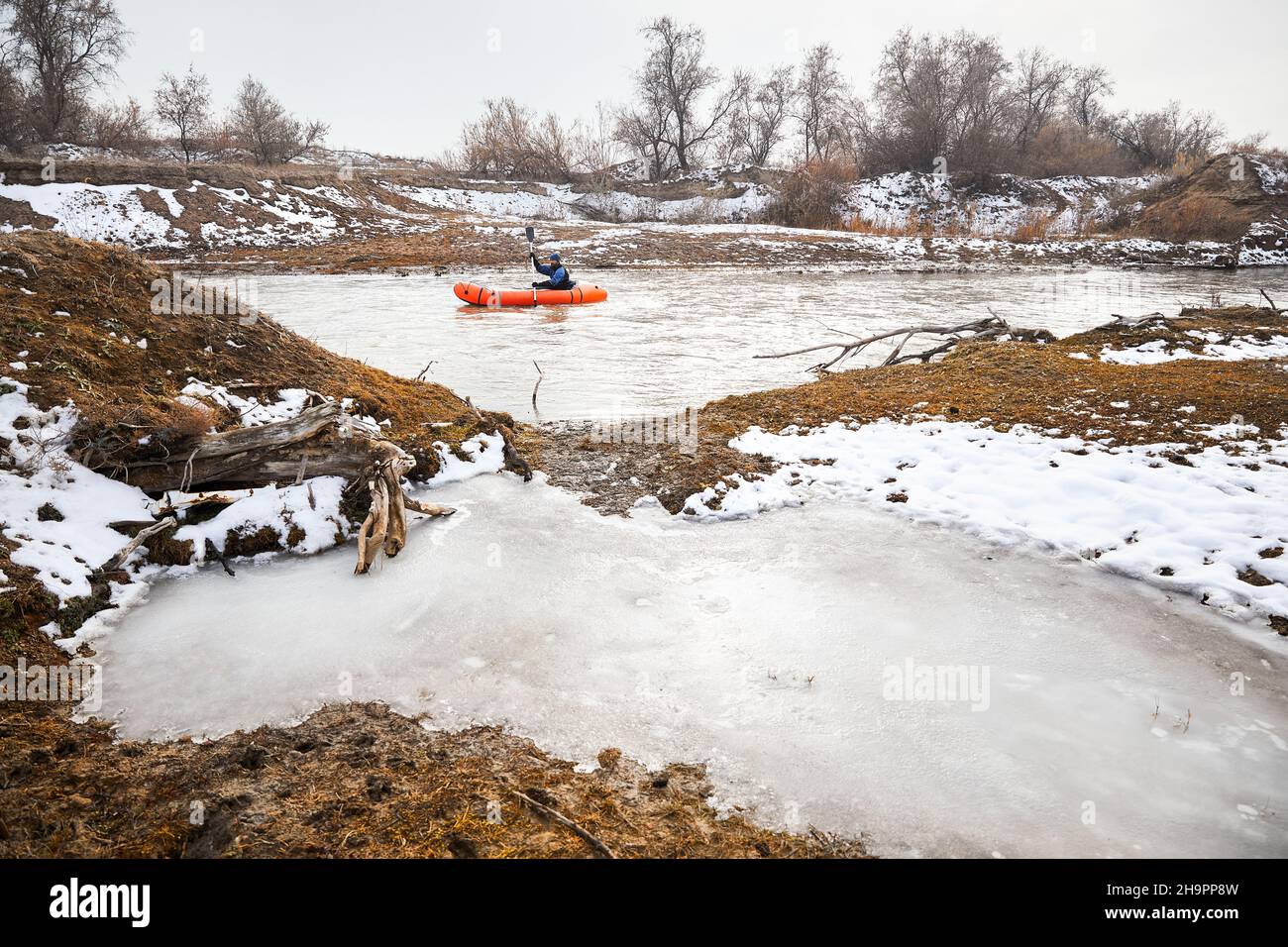 Il vecchio uomo con barba in giacca blu è a pagaiare su una barca sportiva di zattera arancione nel fiume di acqua sporca in inverno con la neve banca. Foto Stock