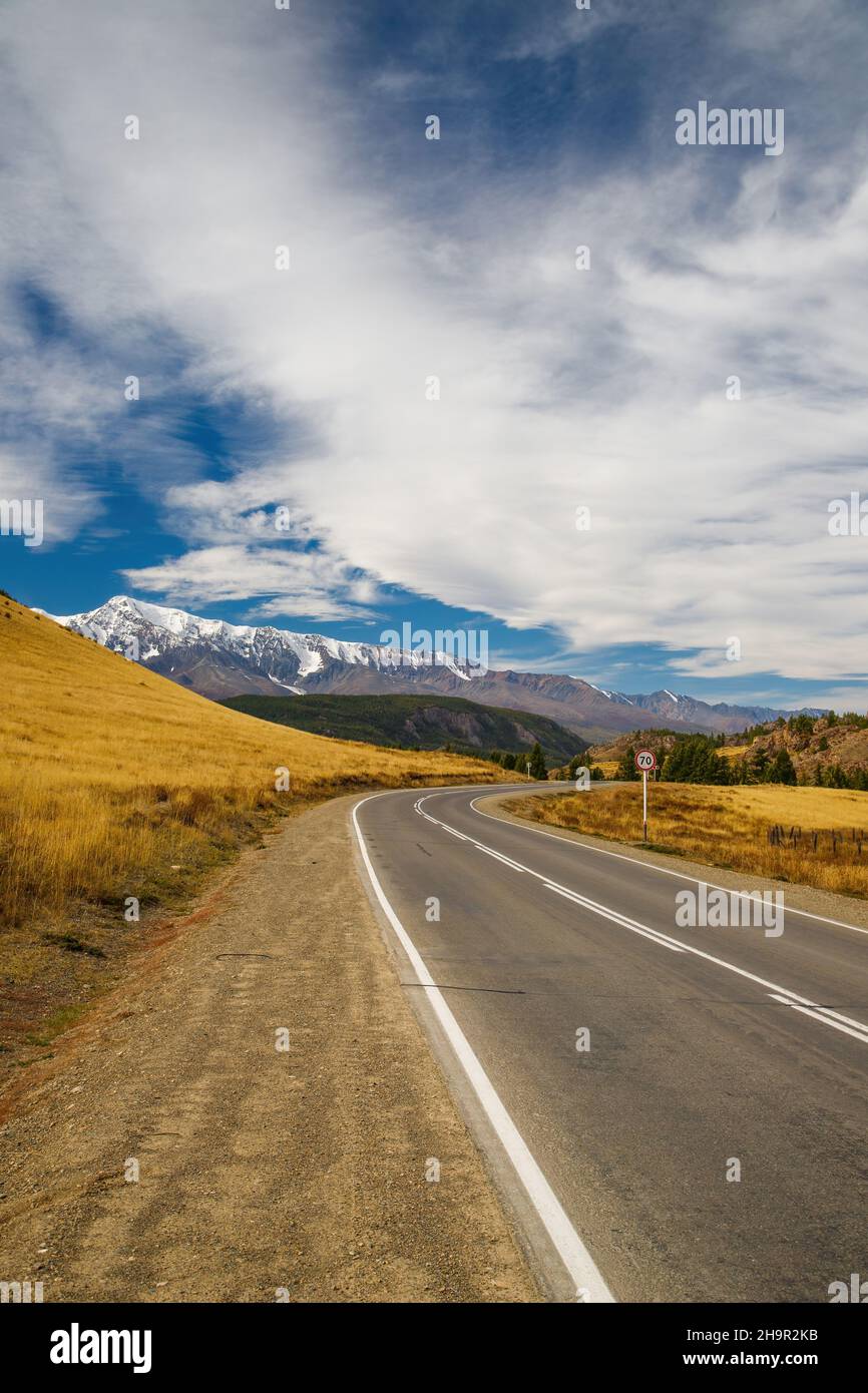 Foto in formato ritratto di un'autostrada vuota e diritta che conduce alle vette innevate della catena montuosa del Kuray. Bel cielo blu nuvoloso come sfondo. Alta Foto Stock