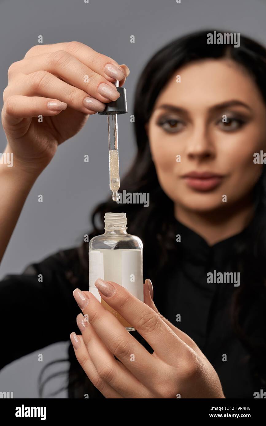 Cosmetologo caucasico femminile in uniforme nera posato su sfondo grigio e contenente siero facciale con pipetta. Cura della pelle e trattamento concetto. Foto Stock