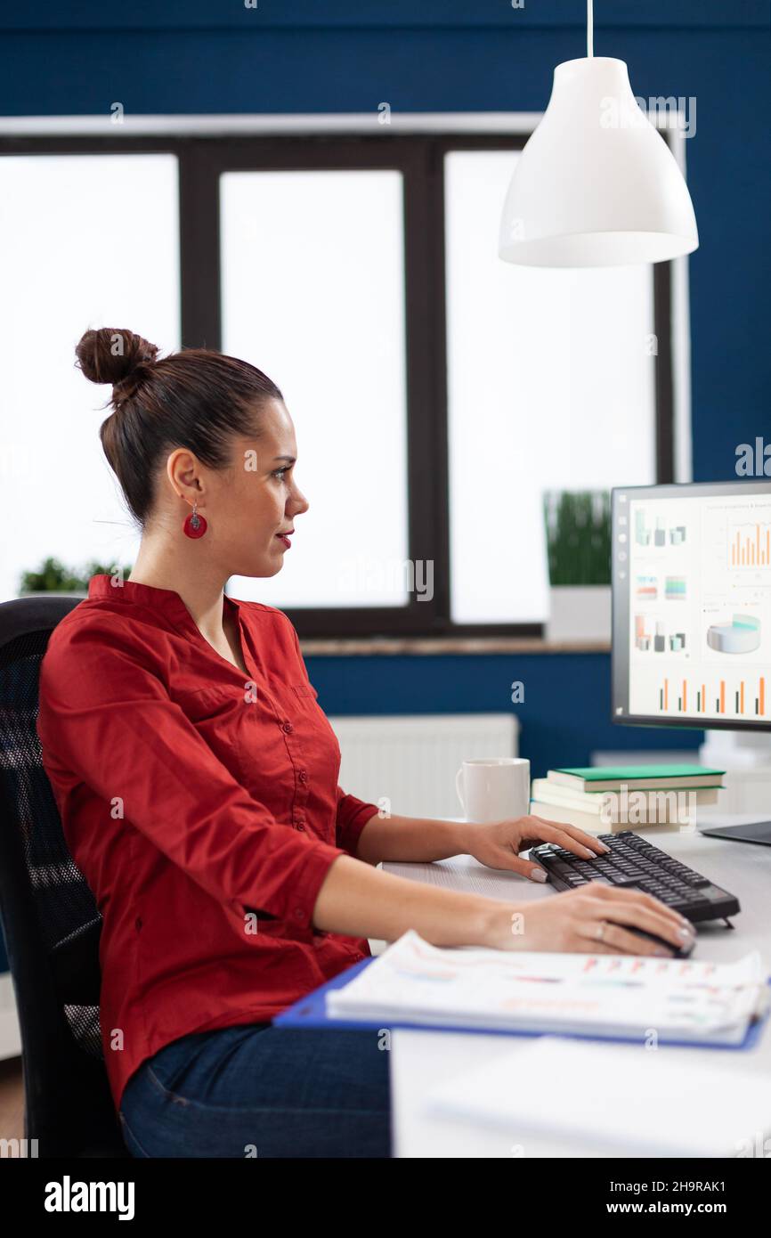 Proprietario di avvio con appunti che lavorano alla scrivania con lampada da soffitto. Imprenditore che analizza business chart sullo schermo del computer. Donna d'affari in camicia rossa digitando i dati sulla tastiera. Foto Stock