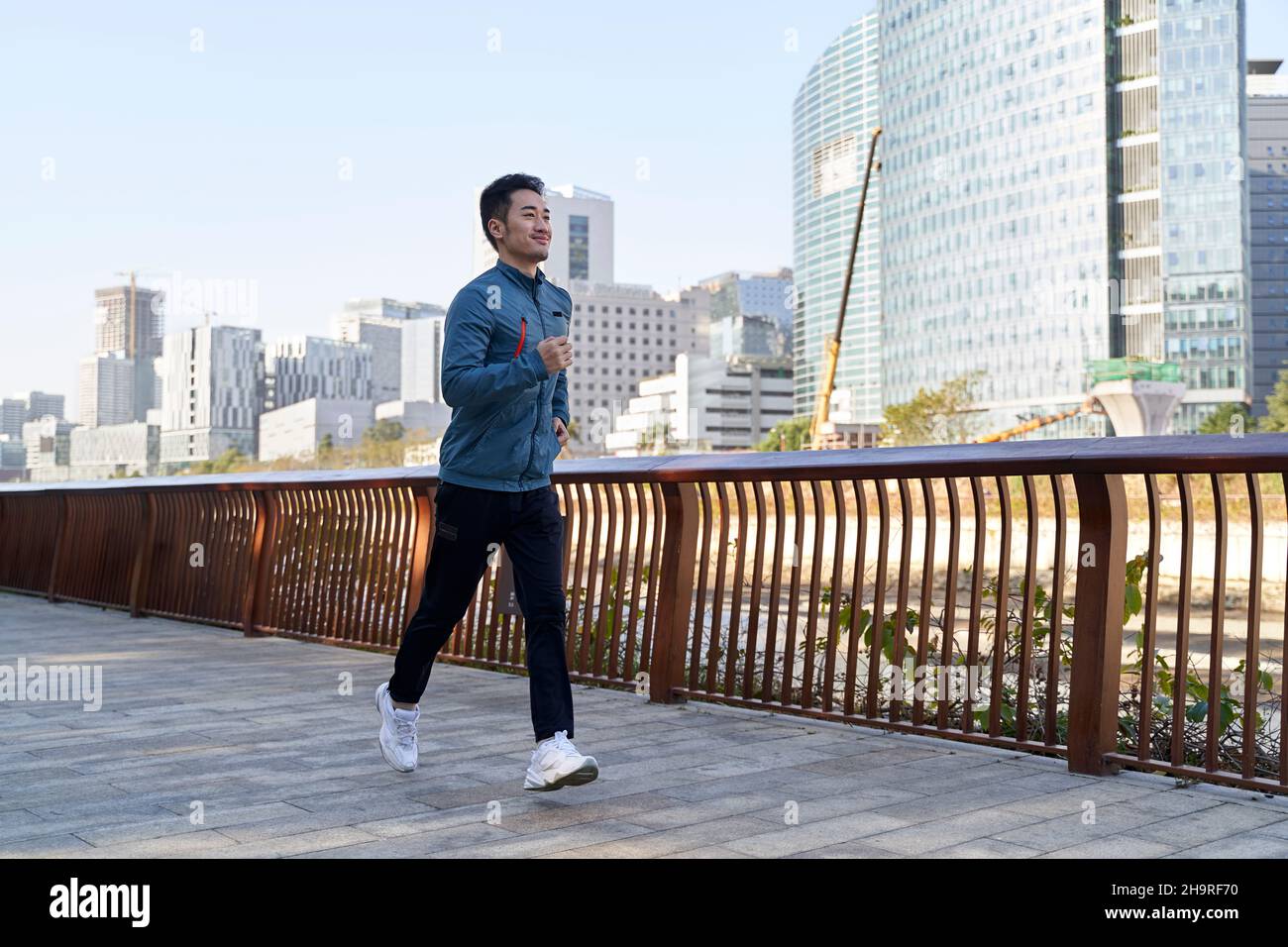 giovane uomo asiatico che corre jogging su un sentiero lungofiume nel parco pubblico con paesaggio urbano sullo sfondo Foto Stock