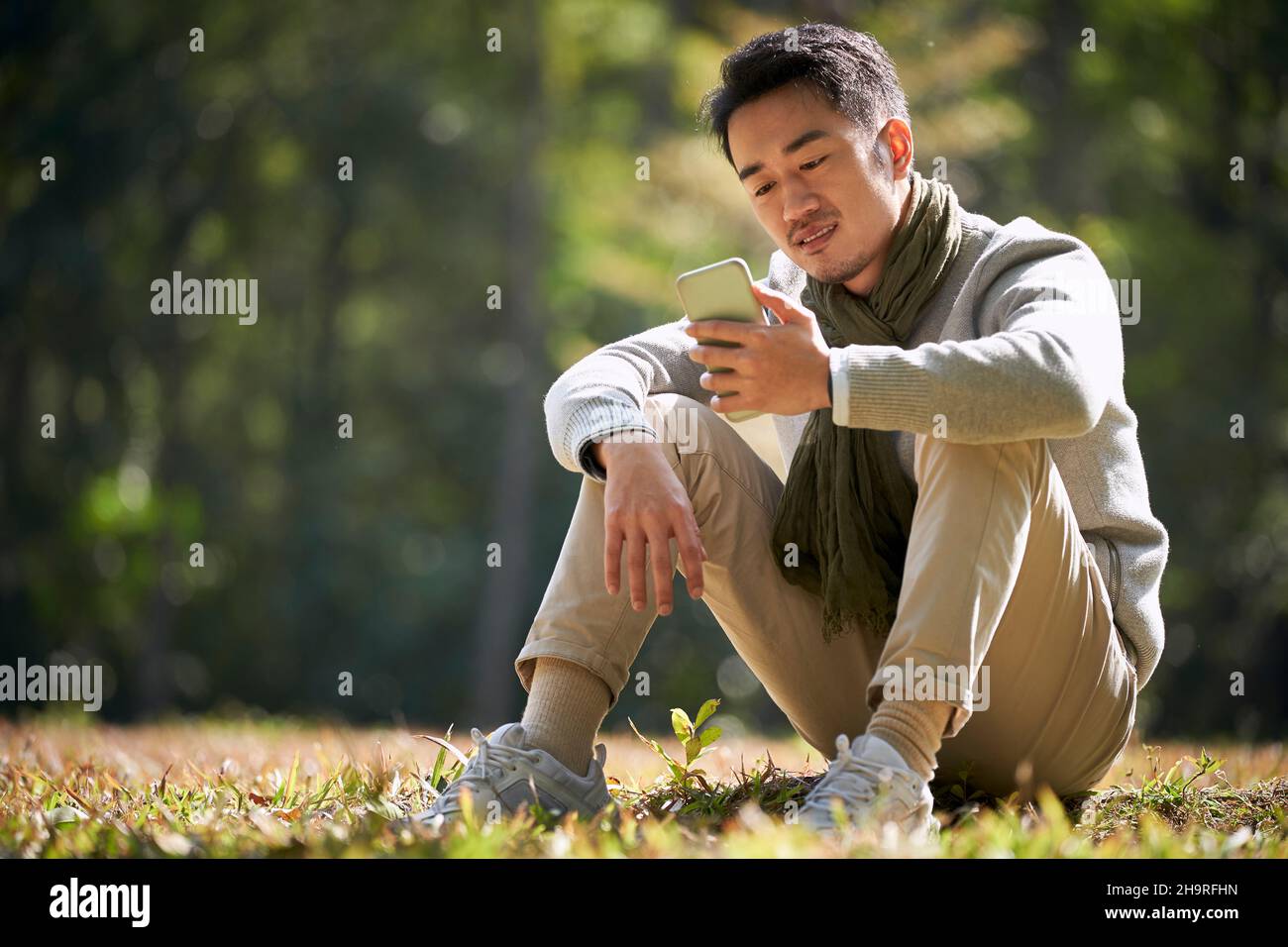 giovane uomo asiatico seduto in erba guardando il cellulare all'aperto nel parco della città Foto Stock