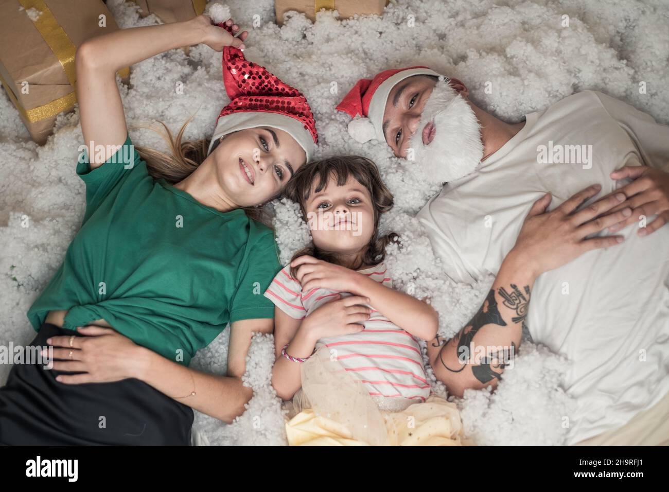 Foto di Natale di Happy Family. Capodanno Ritratto di papà, mamma e figlia con cappelli rossi di Natale, sorridente e posato in neve artificiale bianca. Foto Stock