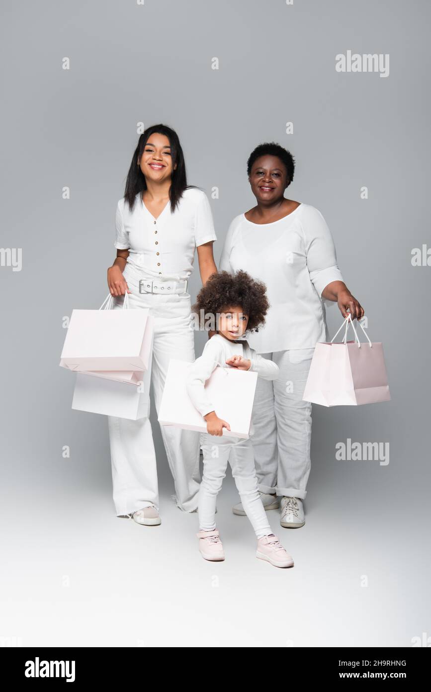 sorridente capretto afroamericano con madre e nonna che tiene i sacchetti bianchi dello shopping sul grigio Foto Stock