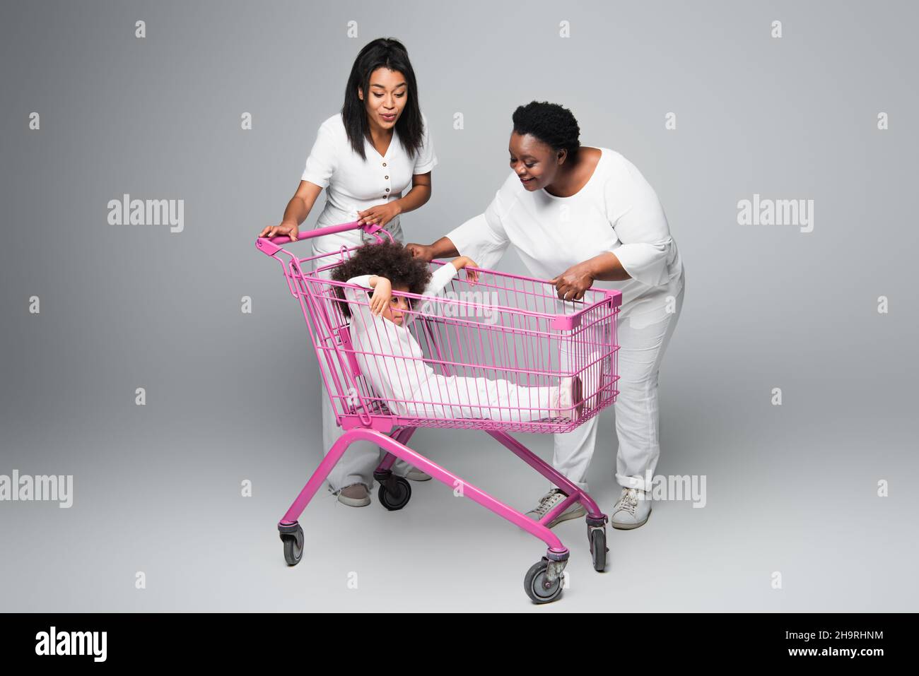 ragazza afroamericana seduta nel carrello dello shopping vicino a mamma felice e nonna sul grigio Foto Stock