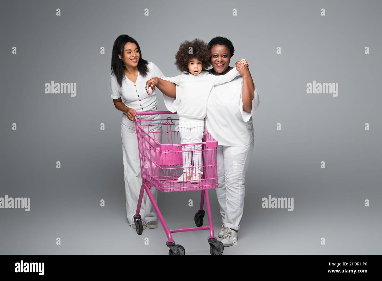 curly african american ragazza in piedi nel carrello vicino a mamma felice e nonna in grigio Foto Stock
