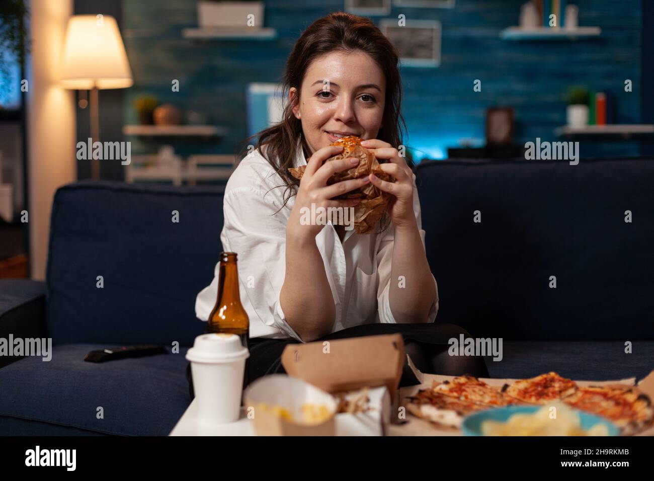 Donna sorridente che tiene in mano gustosi hamburger guardando la televisione commedia sitcom in piedi sul divano di fronte al tavolo con menu fast food takeaway. Happy person guardando lo spettacolo televisivo che ha takeout cena. Foto Stock