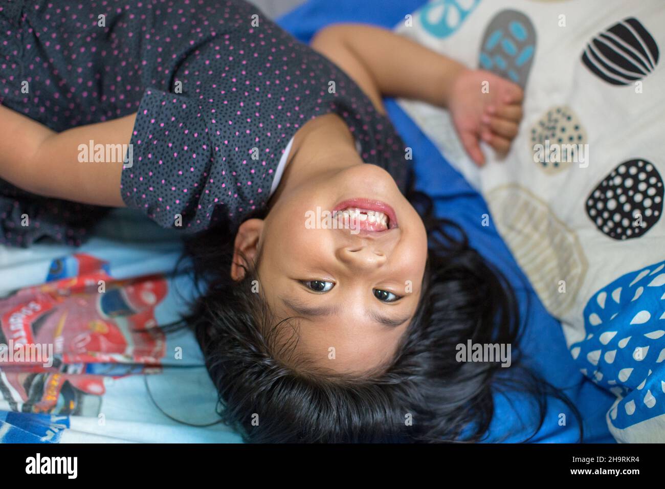 Una bella bambina con lunghi capelli neri con denti mancanti sorridenti sul letto, sparare da un angolo alto Foto Stock