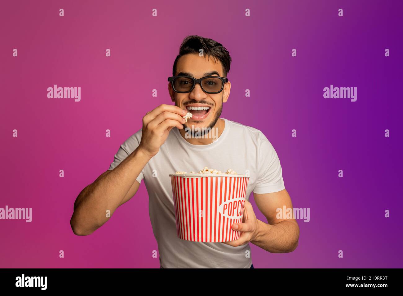 Ritratto di felice ragazzo arabo giovane in 3D bicchieri mangiare popcorn dal secchio, guardare film al cinema, neon luce Foto Stock