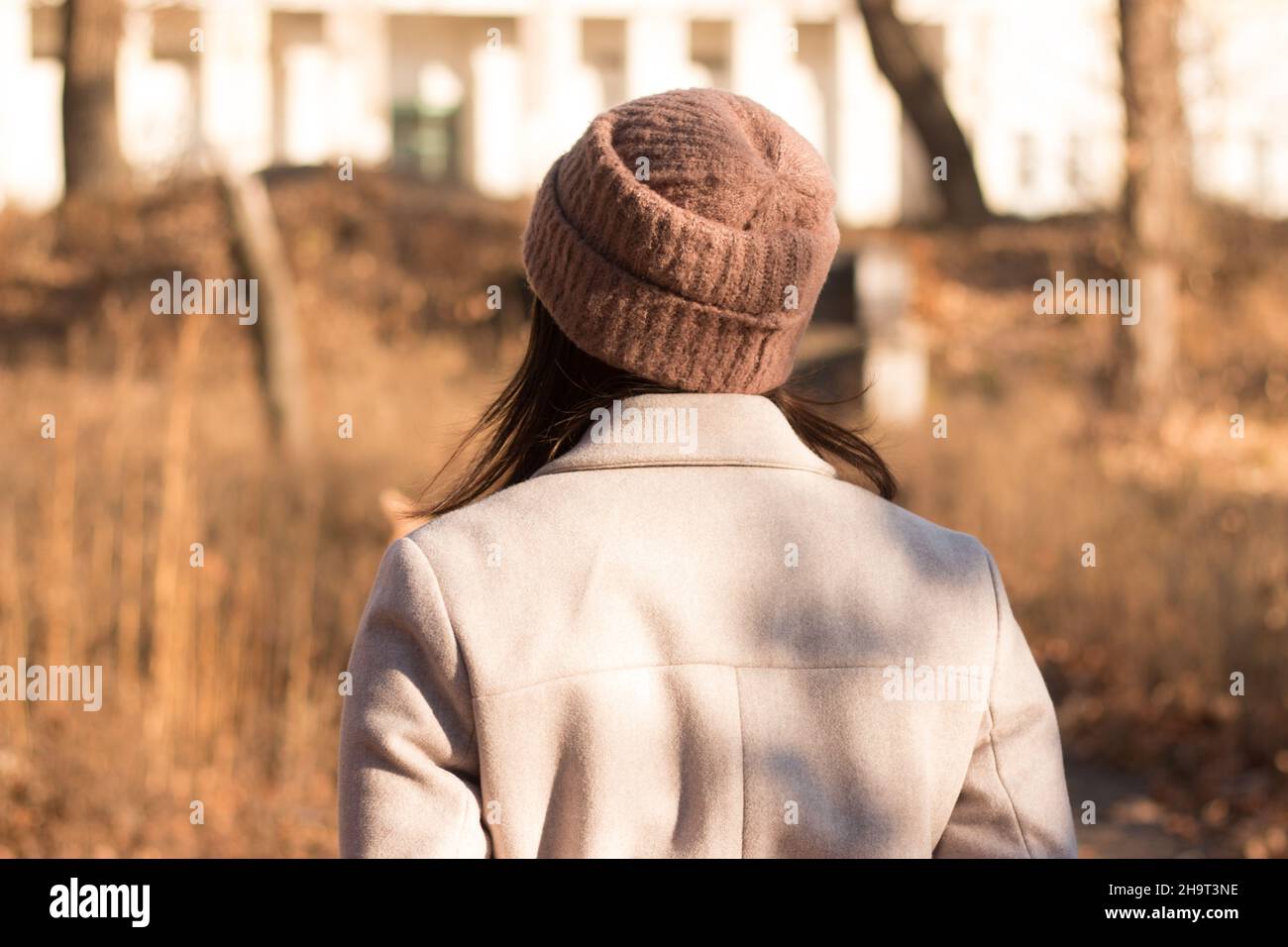 Una ragazza con lunghi capelli scuri in un cappotto e un cappello a maglia  si alza con la schiena alla macchina fotografica sullo sfondo di un vuoto  parco autunnale Foto stock -