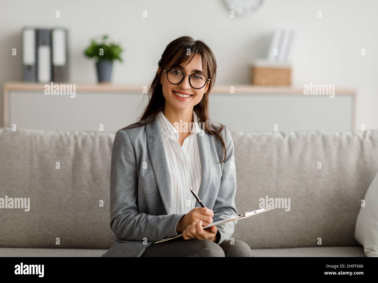 Concetto di terapista professionista. Ritratto di psicologo arabo femminile seduto sul divano, tenendo appunti e sorridente Foto Stock