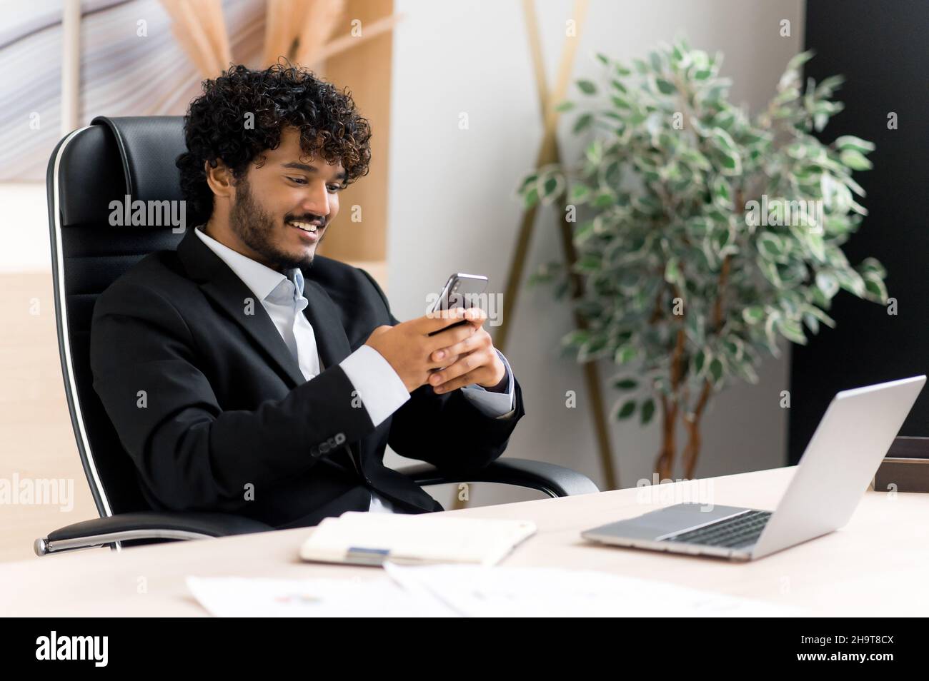 Comunicazione remota tramite smartphone. L'uomo positivo indiano seduto alla sua scrivania in ufficio, inviare messaggi di testo online il suo gadget, risponde e-mail, naviga in internet, sorridendo amichevole Foto Stock