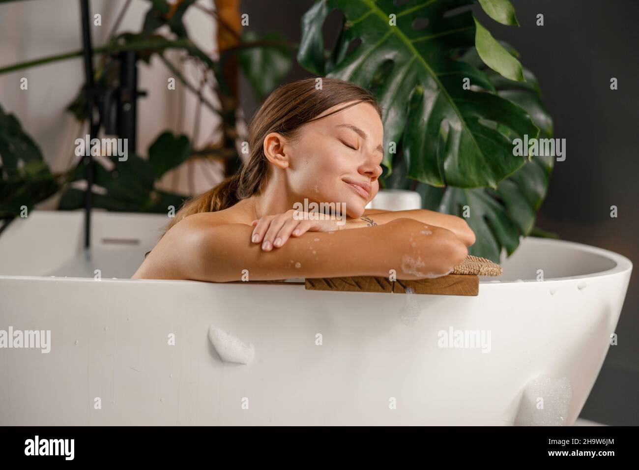 Giovane donna sognante appoggiata sul lato della vasca da bagno e riposata nel bagno decorato con piante tropicali Foto Stock