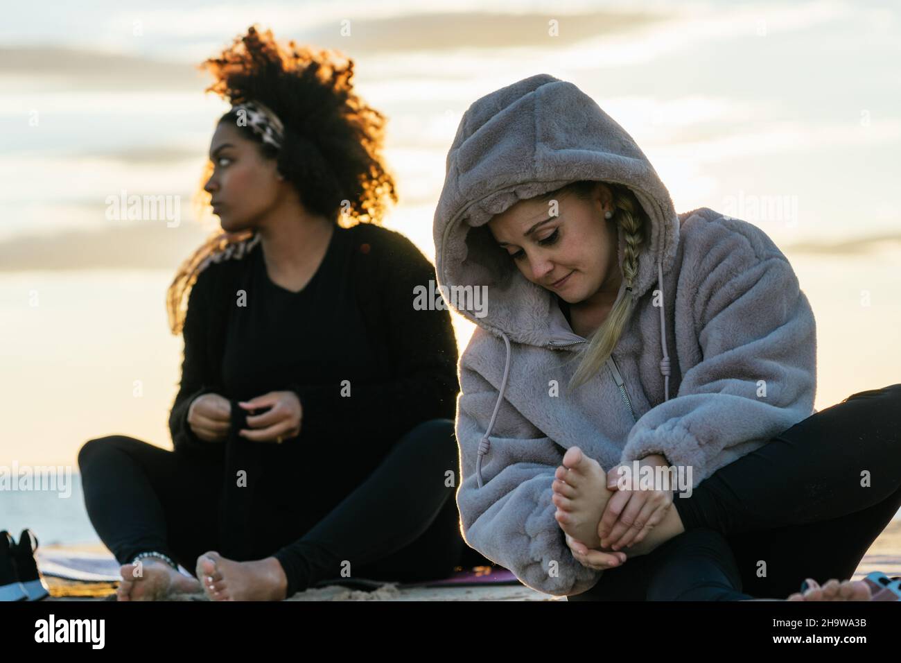 Due donne in abiti caldi seduti su una spiaggia al tramonto dopo una lezione di yoga Foto Stock