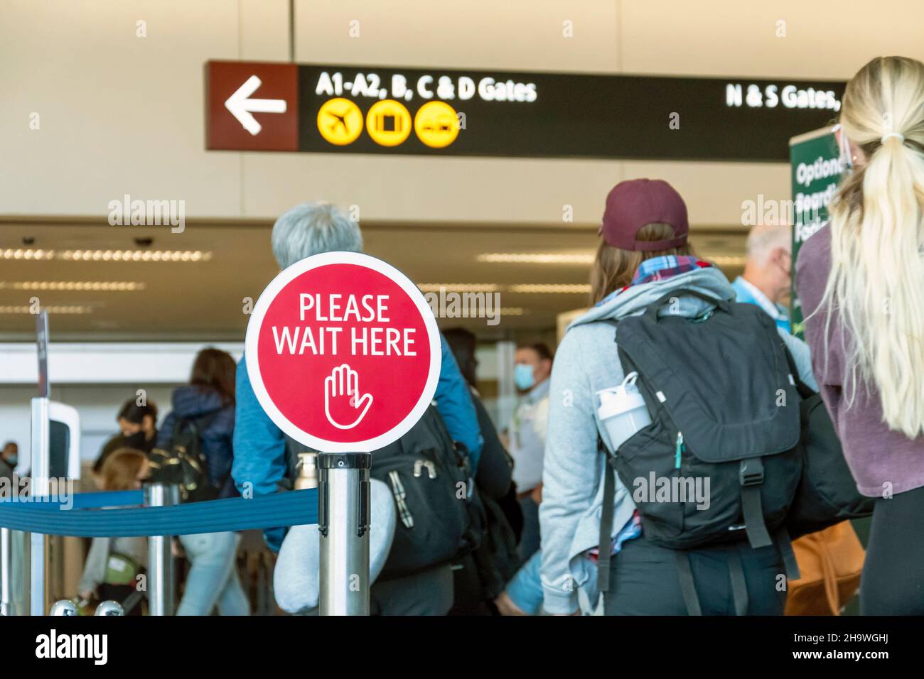 I viaggiatori aspettano in fila in un aeroporto trafficato durante la covid accanto ad un cartello per favore attenda qui. Foto Stock