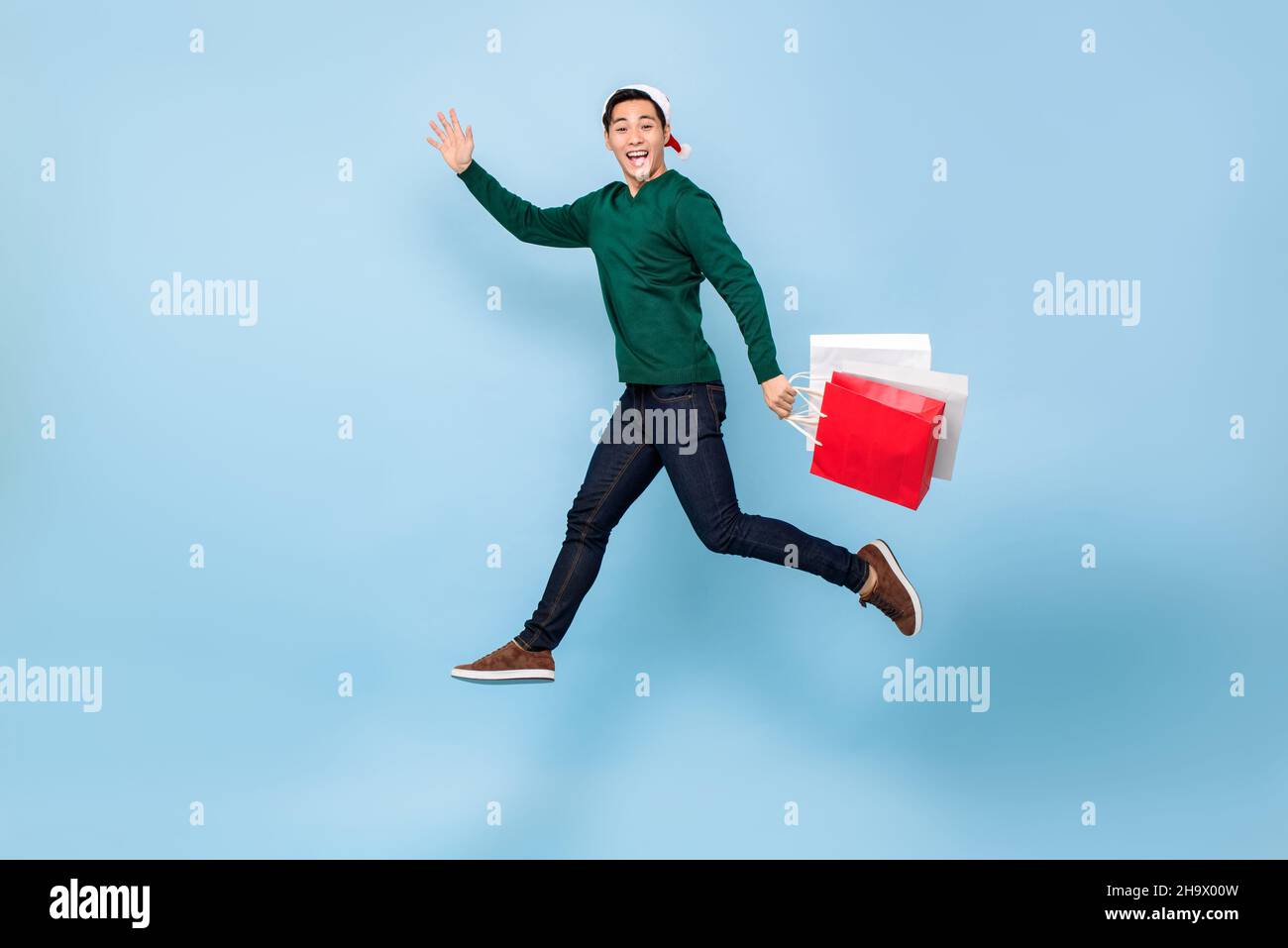 Ritratto a tutta lunghezza del giovane uomo asiatico felice che indossa il cappello di Natale che salta e tiene borse shopping in isolato sfondo azzurro studio Foto Stock