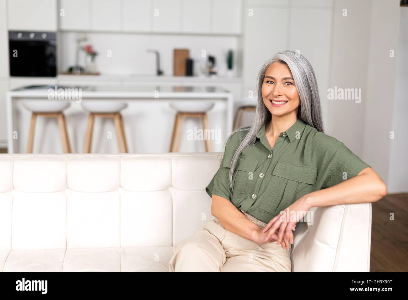 Ritratto di attraente donna di mezza età con lunghi capelli d'argento seduta in appartamento moderno, metà di età multirazziale asiatico donna riposo a casa, guardando la macchina fotografica e sorridendo con ampio sorriso toothy Foto Stock