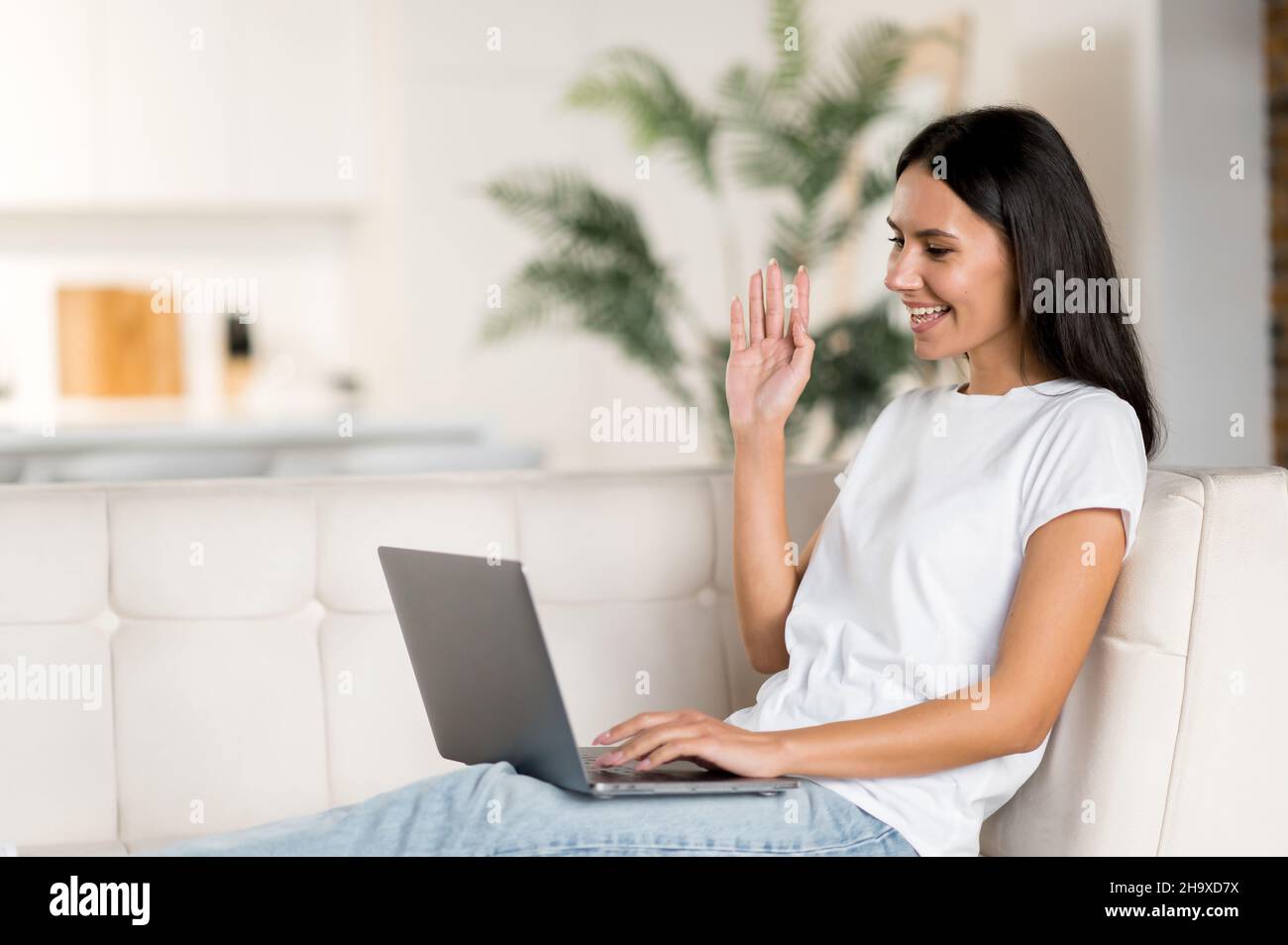 La giovane donna caucasica felice usa il laptop, facendo una videochiamata, comunica con i colleghi o gli amici in videoconferenza, agitando la mano e sorridendo. Bella donna brunetta si siede su un divano in soggiorno Foto Stock