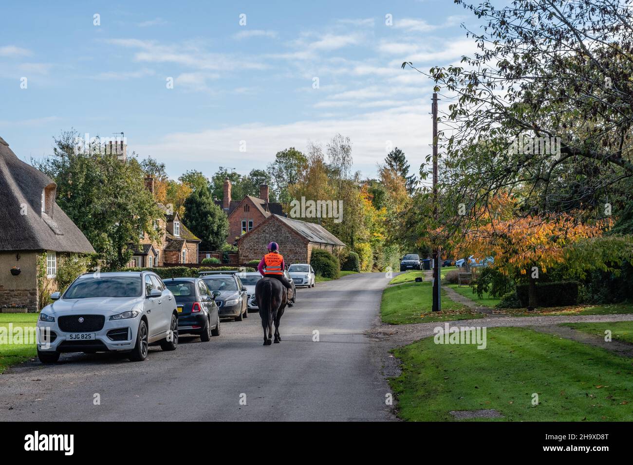 Scena stradale, con case di paglia, nel grazioso villaggio di Ashby St Ledgers, Northamptonshire, Regno Unito; cavaliere nel centro della strada Foto Stock