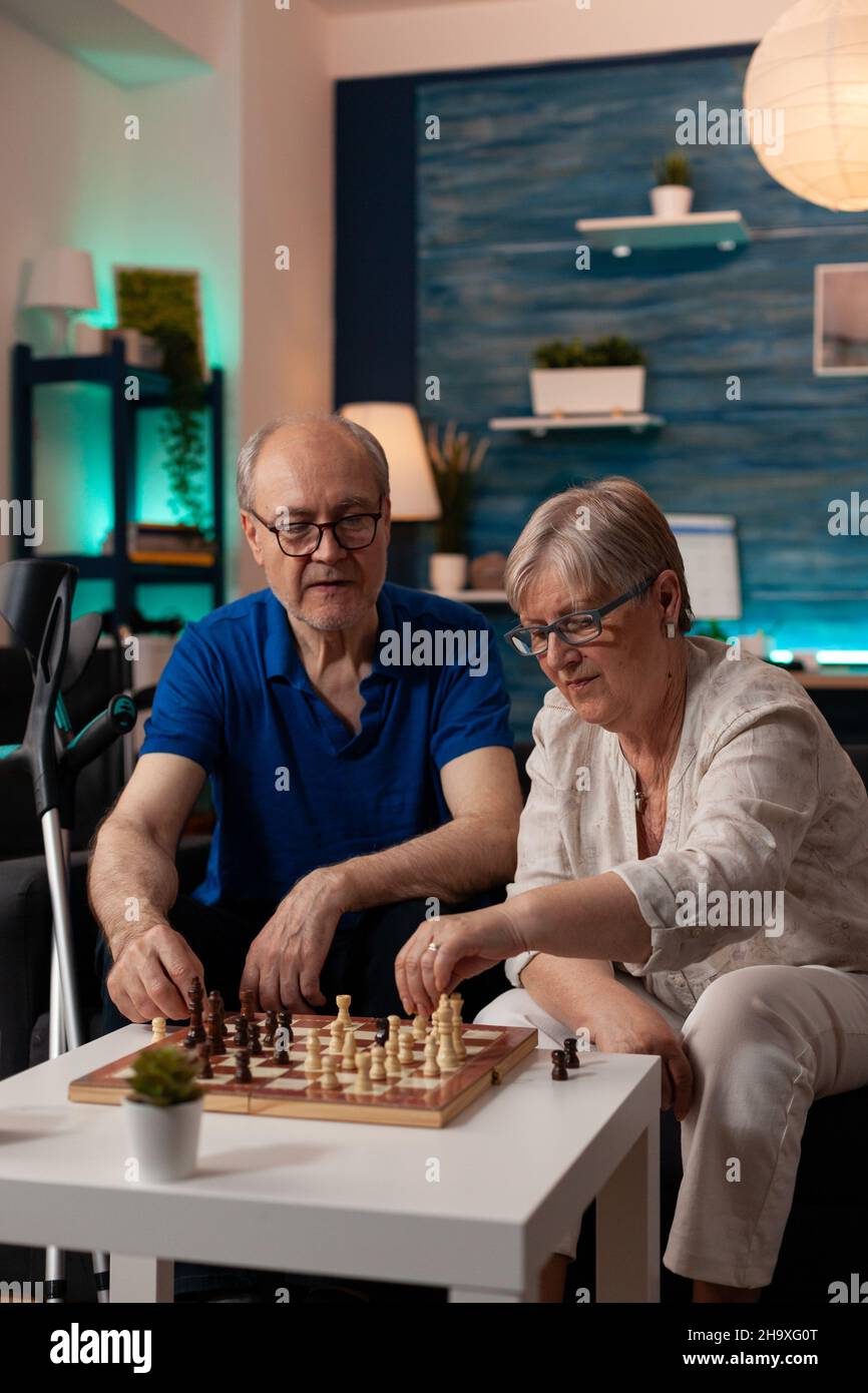 Persone anziane che godono di pensione con gioco di scacchi sul tavolo da caffè in soggiorno. Uomo e donna anziana seduti sul divano con scacchiera che giocano insieme per divertimento e divertimento a casa Foto Stock