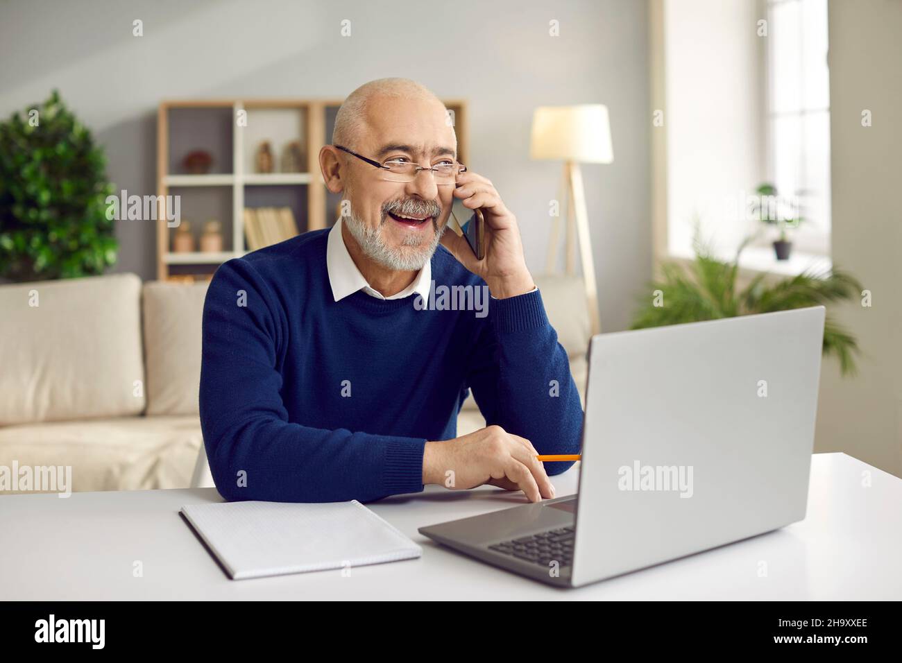 Uomo d'affari anziano che parla sul telefono delle cellule che si siede davanti al laptop nell'ufficio domestico. Foto Stock