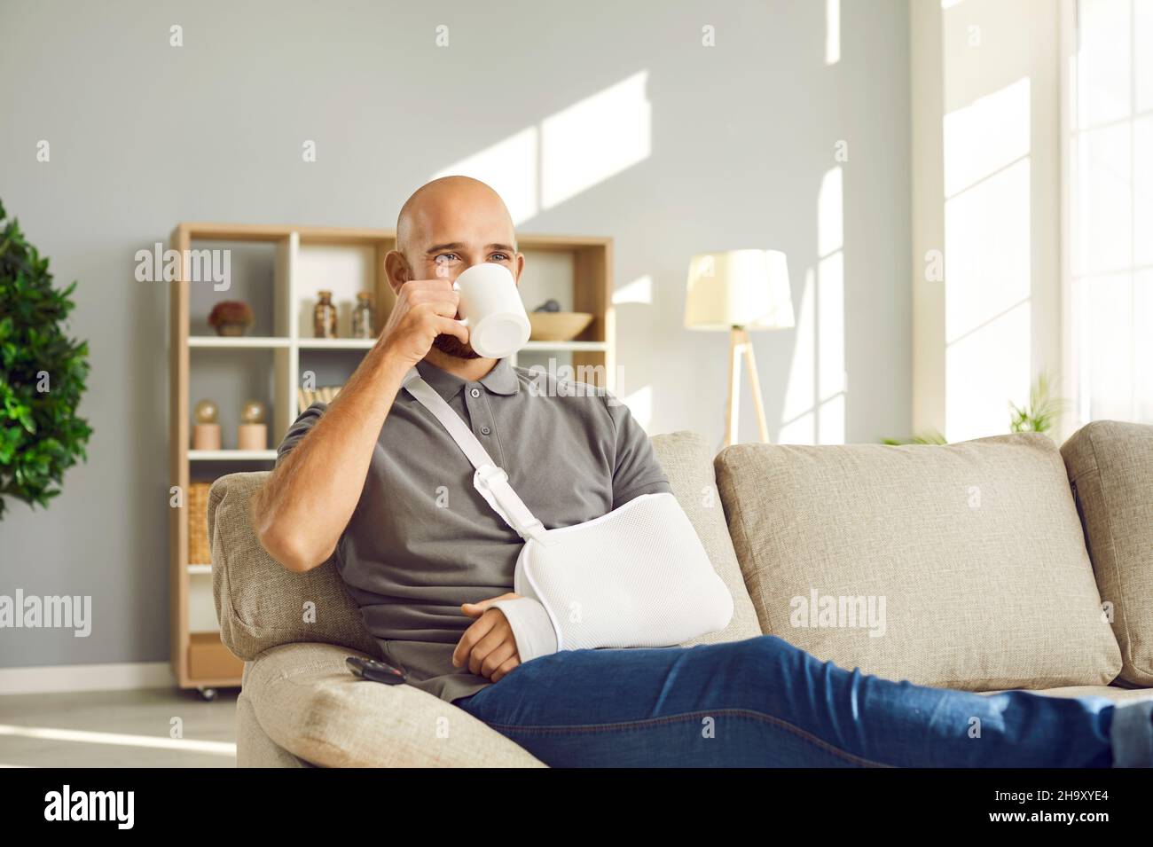Buon giovane uomo con braccio rotto seduto sul divano a casa e una tazza di caffè Foto Stock