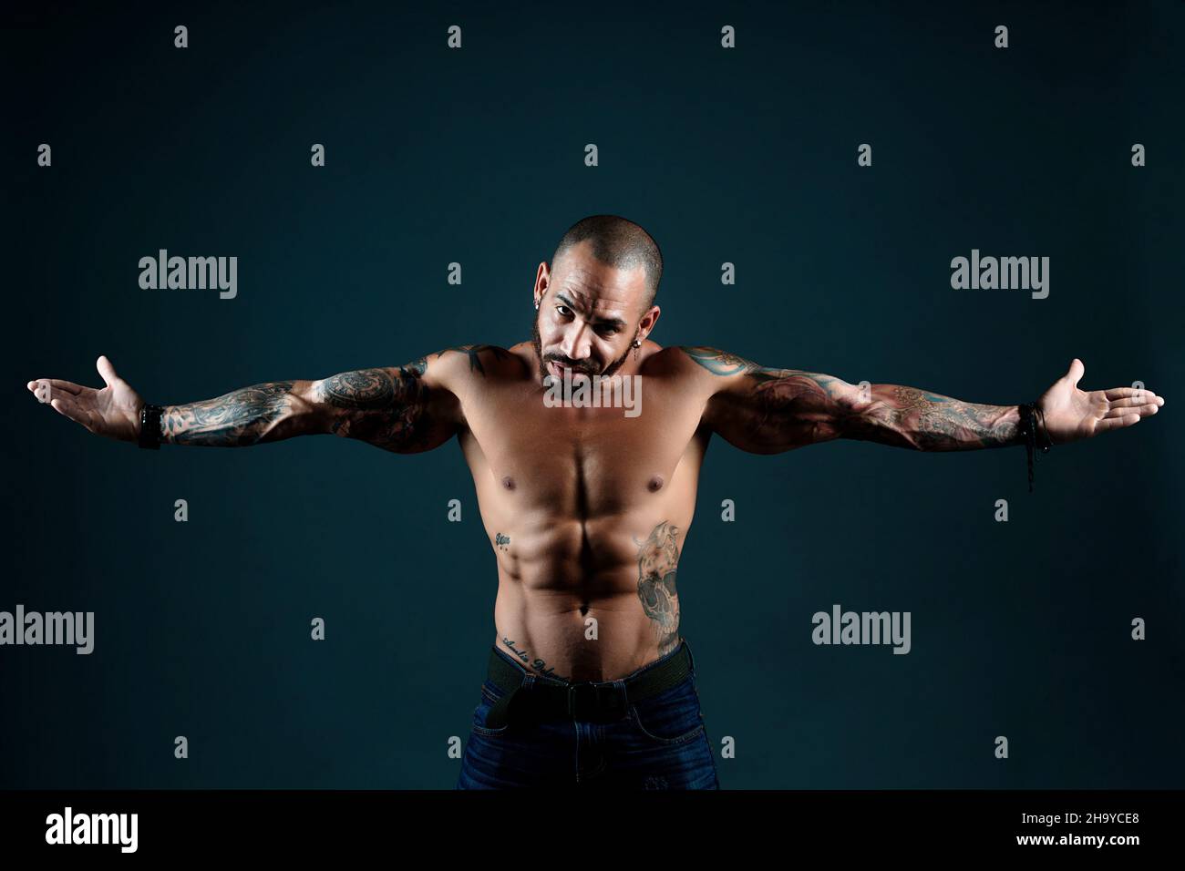 Il muscoloso Latino o Cubano con tatuaggi sul corpo si pone in studio su sfondo scuro. Gesta e guarda la fotocamera. Uomo sicuro e senza paura con taglio corto. Bandito. Foto Stock